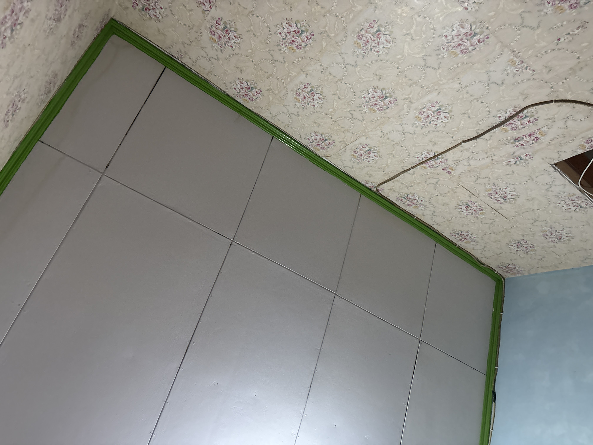 The image shows a ceiling in a room with light gray tiles. A green trim runs along the edge of the ceiling where it meets the walls. The walls are covered in floral wallpaper with a cream background and pink and white flowers. Electrical wires are visible on the wall, running along the wallpaper. One wall appears to be painted light blue. The scene suggests an interior residential or commercial space in Palimanan, Indonesia.