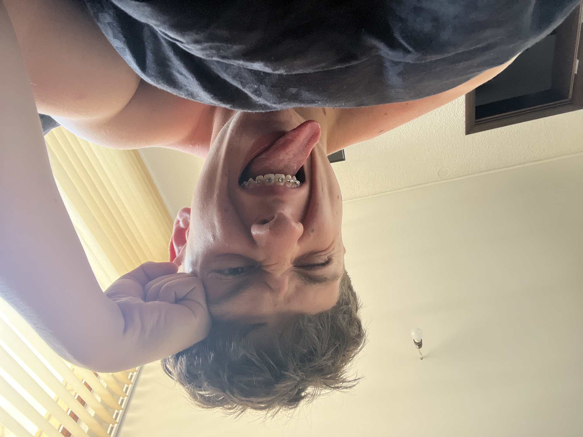 A young person with brown, messy hair and braces is in a close-up selfie, their face contorted in a playful, winking expression with their tongue sticking out. Their right hand is propped against their forehead, and they are partially covered by a dark blanket. The background shows a white wall with a light fixture and a door, and in the upper right corner, there are vertical blinds. The overall lighting suggests an indoor setting, possibly a bedroom.