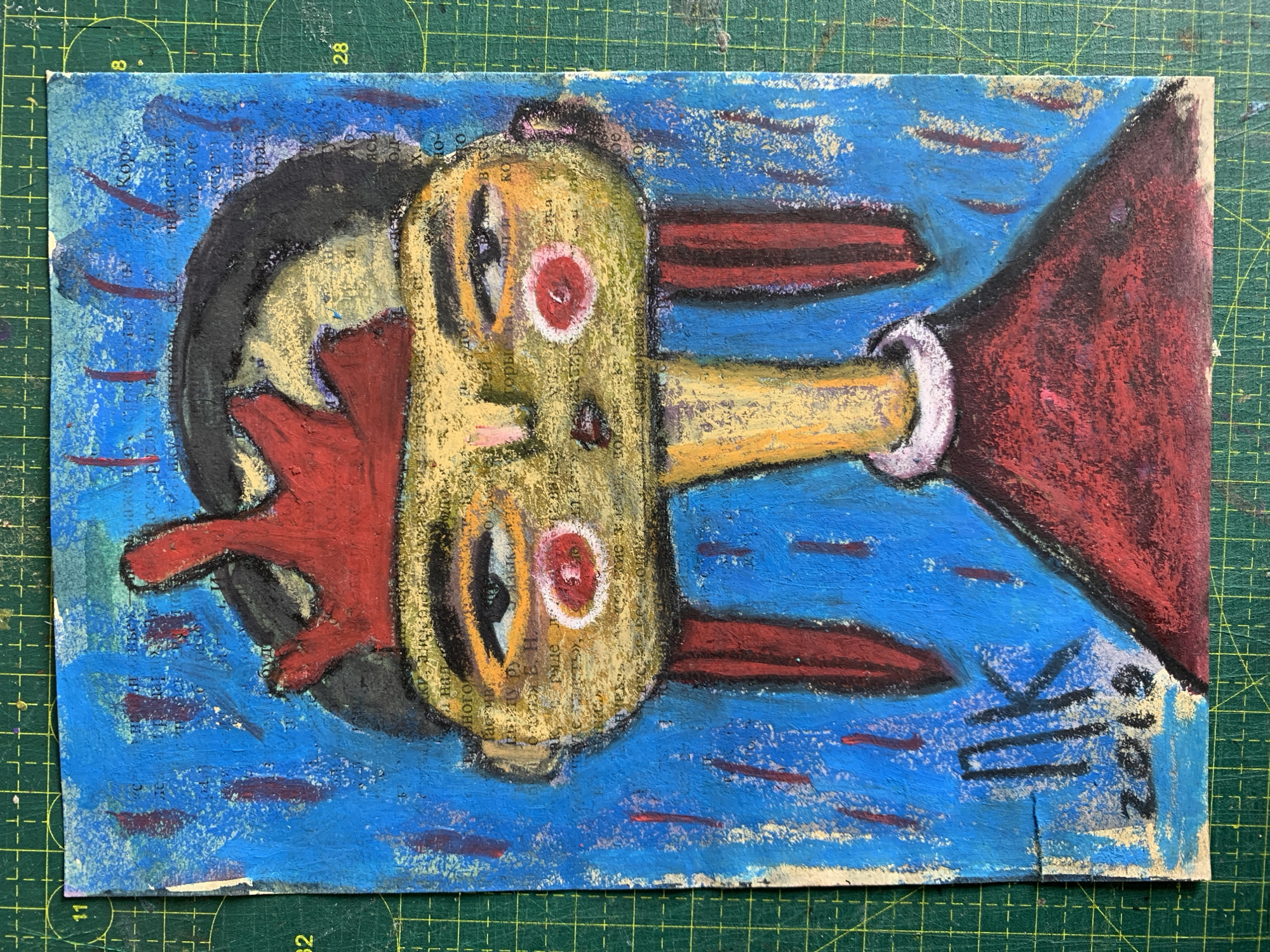 The image is a portrait of a stylized figure with a red crown, drawn on paper that appears to be collaged with text from a book. The background is a vibrant blue with red vertical marks. The figure has a large head with prominent eyes, cheeks painted red, and what appear to be red hair extensions or adornments. The body is depicted in a simple red garment. In the bottom left corner, there is a signature "NK" and the year "2019". The artwork is placed on a green cutting mat with grid lines, suggesting it is an artist's workspace. There is no information within the image that provides any context about the city of Sergeevo, Russia.