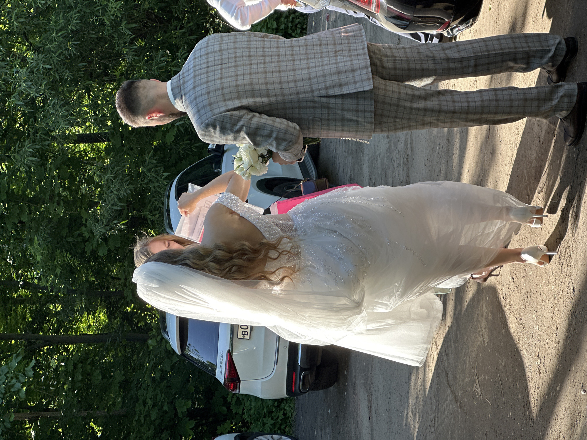 The image depicts an outdoor wedding scene under bright sunlight. In the foreground, a woman, presumably the bride, stands with her back partially towards the viewer. She wears an embellished white wedding gown with a low back, a flowing veil, and white high-heeled shoes. Her long, wavy blonde hair is visible beneath the veil. She holds and appears to be examining some papers or documents in her hands.
To her right, a man, likely the groom, stands facing generally towards the bride. He is dressed in a grey and brown plaid suit, consisting of a jacket and trousers, and black dress shoes. He holds a bouquet of white flowers in his left hand. Another person, wearing a white long-sleeved top, is partially visible in the upper left background.
The setting features dense green foliage and trees on the left side of the frame, contrasting with a sunlit, paved surface (road or path) on the right. Strong shadows are cast on the ground, indicating clear, sunny weather, possibly during late morning or afternoon.
A white SUV is parked behind the bride, identified by the visible text "GLE 300 D 4Matic" on its rear. Part of its license plate is visible, showing "B0" followed by partially obscured characters and then "80". A portion of another vehicle's wheel is also visible in the bottom left corner. The scene captures a moment during a wedding event, possibly during a pause or preparation.