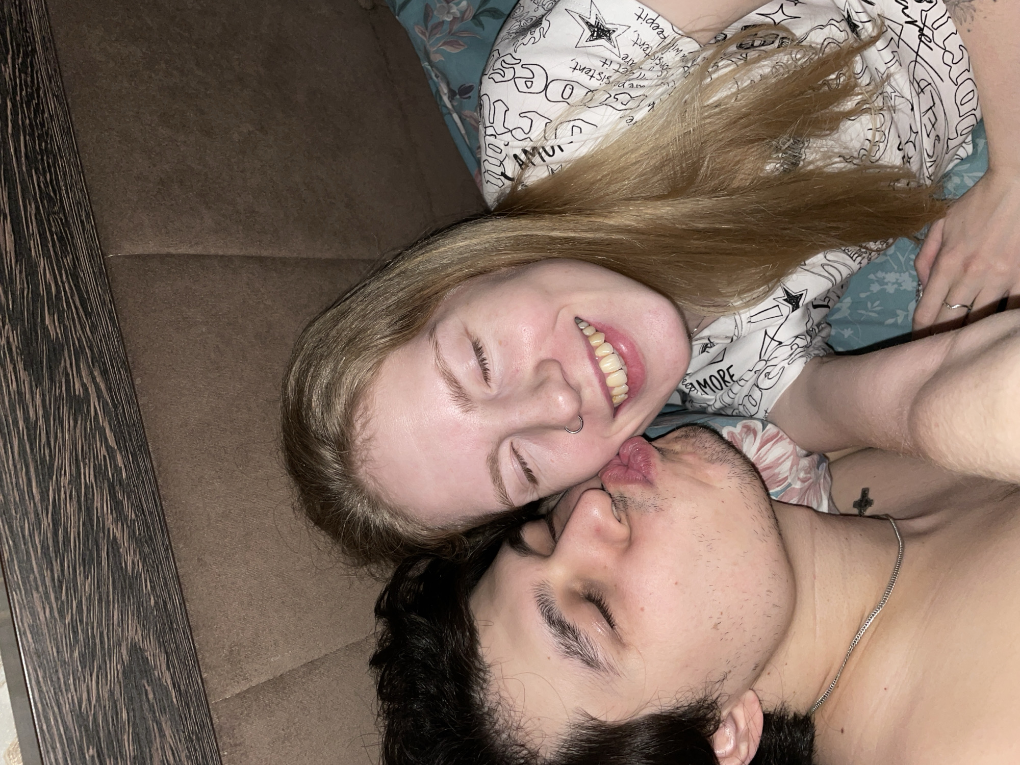 Two people, a light-skinned woman with blonde hair and a light-skinned man with dark hair, are lying down with their faces close together. The woman is smiling and has her eyes closed, and a nose ring is visible. The man also has his eyes closed and appears to be kissing or about to kiss the woman. The woman is wearing a white top with black text and designs, and the man is wearing a silver chain necklace. They are lying on a brown surface that appears to be a couch or bed. Some printed fabric is visible under the woman's head. The image does not contain any information related to Izhevsk, Russia.