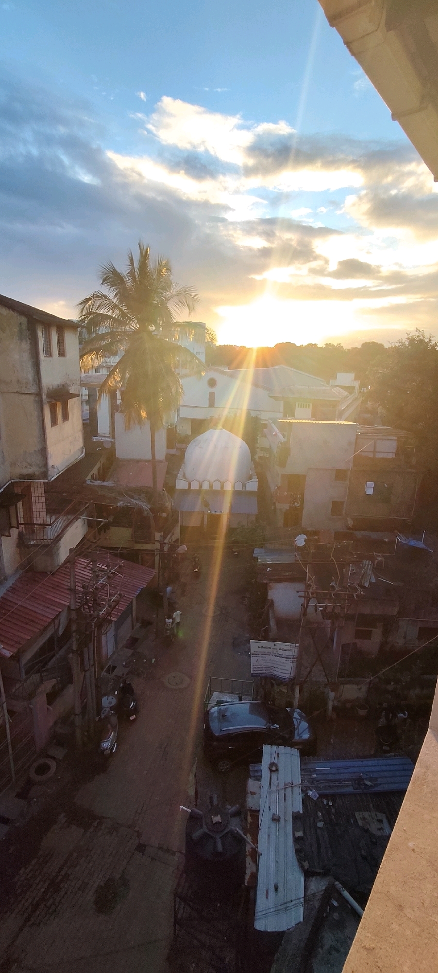 An aerial, vertical, low-angle shot captures an urban scene in Belagavi, India, at sunset. The sun, a bright orb near the center of the frame, casts strong, visible rays through a partly cloudy, light blue sky. The sky transitions to warmer hues of orange and yellow near the horizon.
The cityscape below is densely packed with buildings of varying heights and architectural styles, predominantly residential and commercial. A prominent white dome structure, possibly a mosque or shrine, is visible towards the middle ground. A mature palm tree with a slender trunk and fronds stands tall to the left of the dome, its leaves silhouetted against the bright sky.
A narrow street or alleyway runs through the scene, wet and reflective, suggesting recent rain or the end of monsoon season. Several motorcycles and a dark-colored car are parked or moving along the street. Power lines and utility poles crisscross the area, typical of Indian urban infrastructure. Small shops and residential units with red-tiled roofs line the street. People are present, with a few individuals visible on foot or on vehicles, but their details are indistinct due to the distance and lighting. The overall impression is one of a typical Indian urban neighborhood bathed in the dramatic light of a setting sun.
