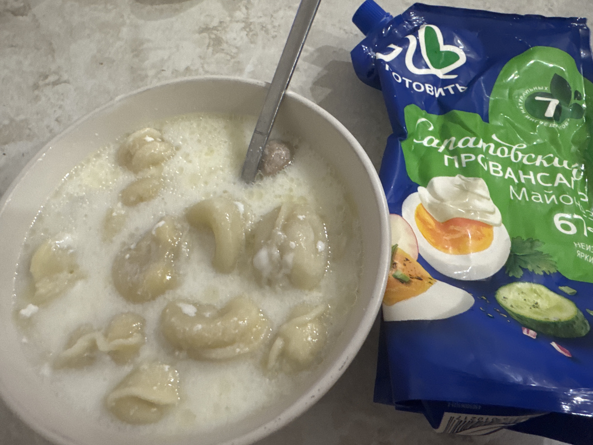 A top-down shot shows a white bowl filled with pelmeni, a type of dumpling, in a creamy sauce. A metal spoon is partially submerged in the bowl. In the foreground, a blue plastic pouch is visible, featuring an image of hard-boiled eggs, cucumber slices, and text in Russian. The scene appears to be indoors, possibly a kitchen, with a textured countertop. The location is specified as Saratov, Russia, based on text on the pouch.