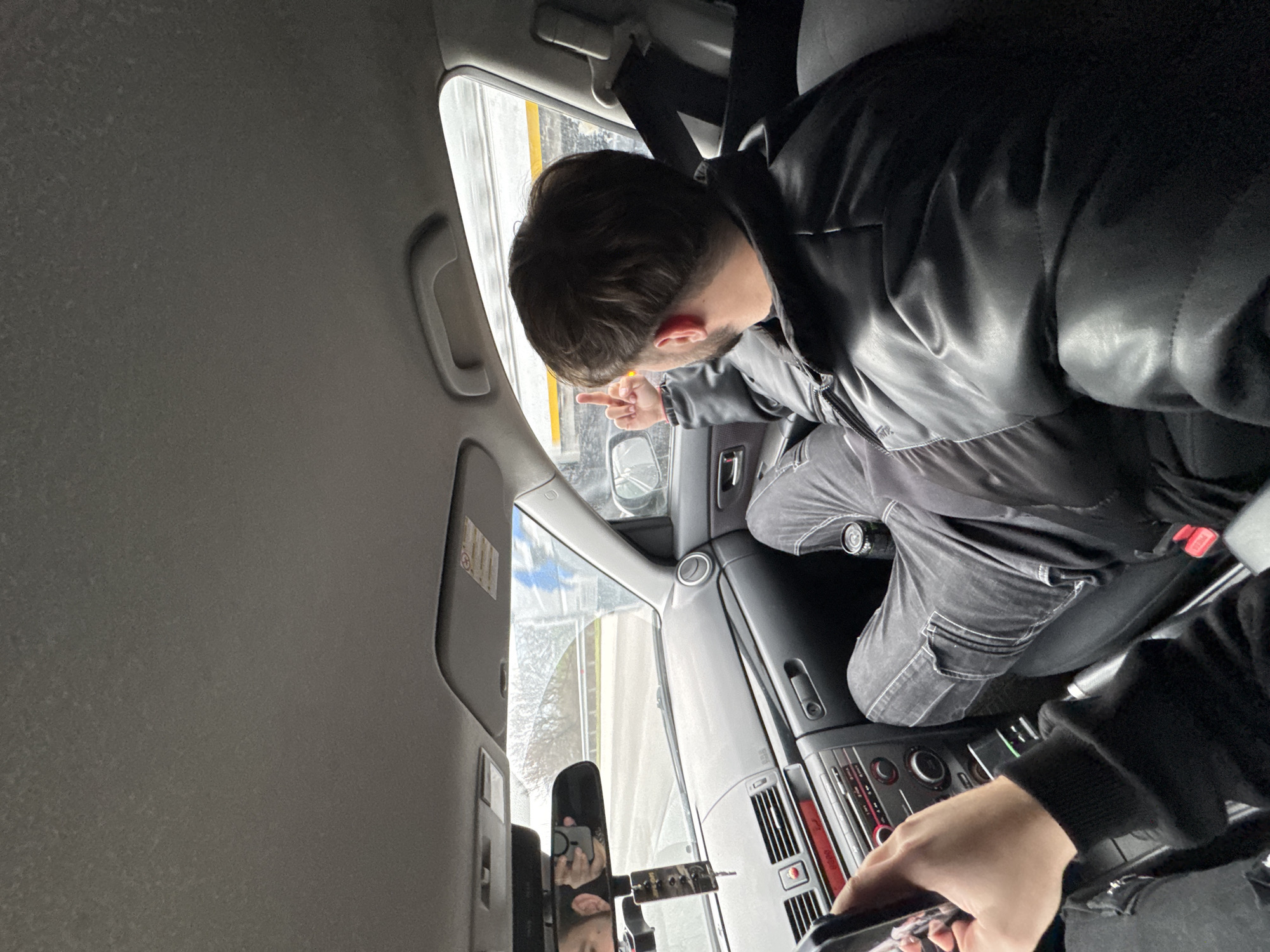 This image captures the interior of a vehicle, specifically the front cabin, in Gabarnitsa, Bulgaria.
A male individual is seated in the front passenger seat, observed from a rear-left perspective. He has short, dark brown hair and a beard, and is wearing a black puffer jacket and dark grey or black jeans. His legs are propped up onto the dashboard. His right arm is raised, with his hand extended and the middle finger prominently displayed towards the vehicle's right side window.
In the foreground, the left hand of another individual, presumably the driver or photographer, is visible, holding a black-cased smartphone. The driver's face is partially reflected in the rearview mirror, along with a dangling car air freshener.
The car's dashboard is dark grey, featuring a radio unit with a red digital display showing "0223" and various buttons and controls. An air vent is visible on the passenger side. A black can, possibly a beverage, rests in a cup holder near the passenger's feet. Through the windshield, a road with a white guardrail and distant vegetation is visible. Through the passenger side window, a blurry white structure with a yellow stripe is discernable. The car's interior ceiling is light grey.
