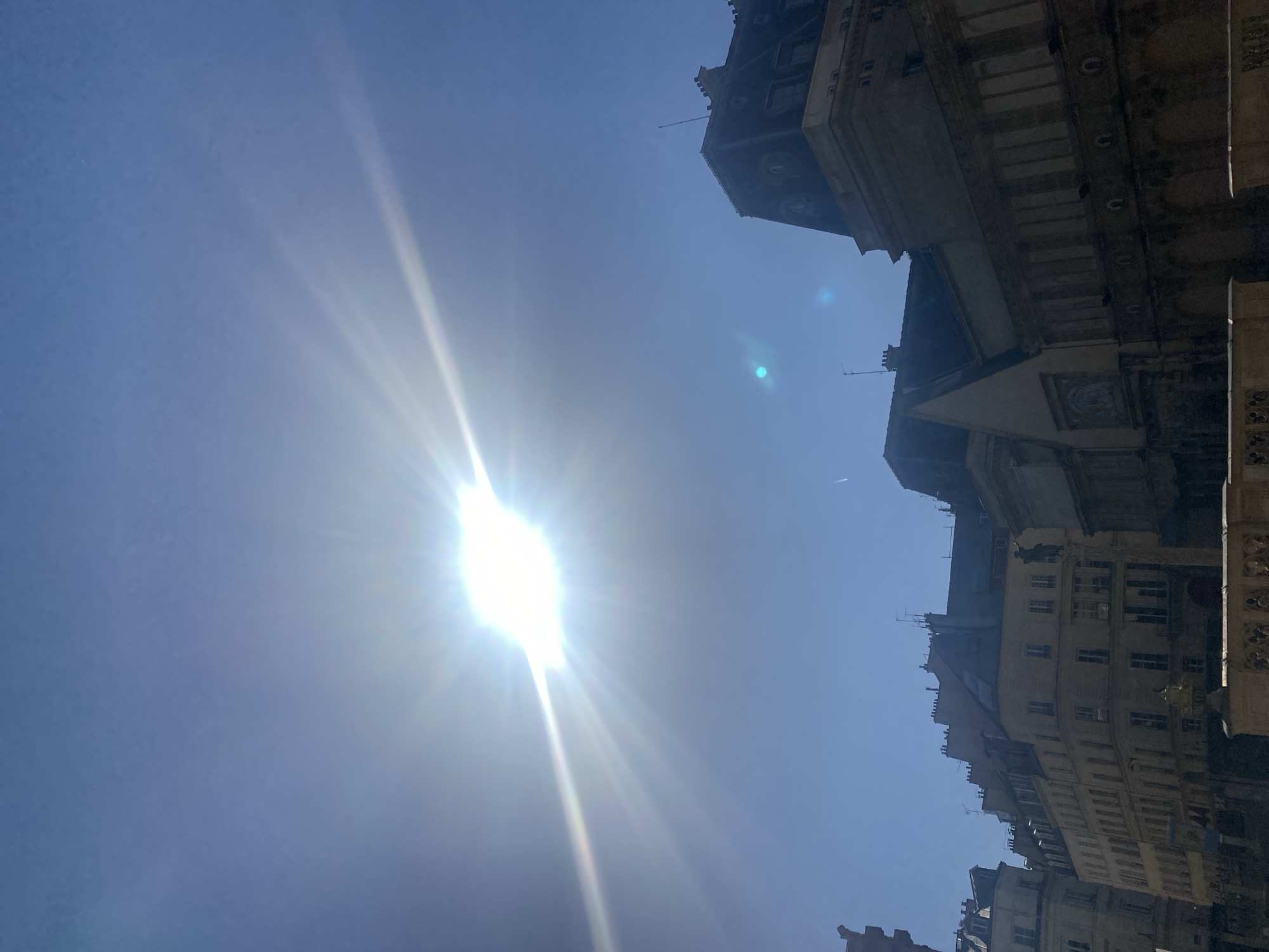 The image depicts a view of buildings in Paris, France, angled upwards towards a bright sun in a blue sky. The buildings are tall, with architectural details including windows, rooftops, and decorative elements. The sun is positioned in the lower left of the frame, creating a strong light source with visible rays.