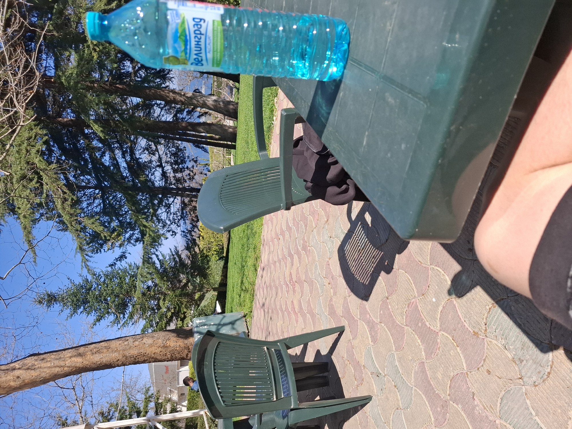 The image shows a green plastic table with a bottle of "Велинград" mineral water on it. There's a green plastic chair at the table, and another similar chair nearby. The ground is paved with patterned tiles. A person's leg is visible next to the table. The background features trees and a blue sky. The scene appears to be outdoors, possibly in a park or garden setting. Without further context, it is challenging to establish that the location is specifically in the city of Varna, Bulgaria, or even to determine with certainty what is the exact location within Varna, without further cues.