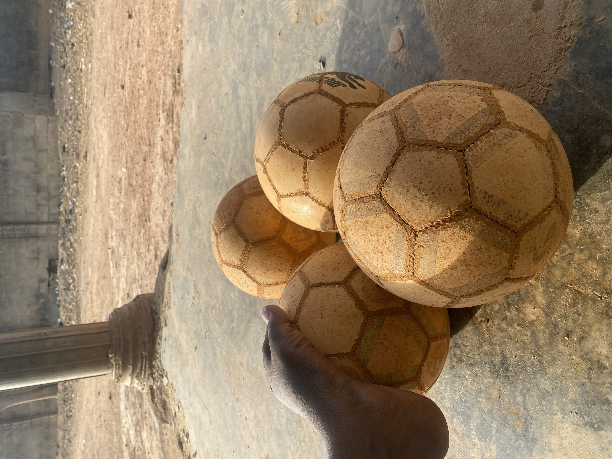 The image displays an outdoor scene featuring four well-used soccer balls resting on a dusty concrete surface. A dark-skinned foot is positioned in the foreground, gently holding or resting against one of the balls.
The four soccer balls are light-colored, possibly white or off-white originally, but are heavily stained and covered in dirt or sand, indicating extensive use, likely on unpaved surfaces. Their classic hexagonal and pentagonal panel patterns are visible, with the stitching lines clearly outlined by accumulated grime. One of the balls, located towards the top center, distinctly shows a visible brand name: "Hunter," accompanied by a stylized logo next to it, which appears to be an abstract or animalistic symbol. The balls show significant wear and tear.
The ground surface is split into two main sections:
1. In the immediate foreground and extending to the right, there is a relatively smooth, grey concrete or paved surface, also speckled with dirt and dust. Strong shadows are cast across this surface, suggesting direct sunlight.
2. To the left and receding into the background, the ground transitions to a sandy or dirt path, covered with loose soil, small debris, and scattered pebbles. This area appears rougher and less maintained.
In the far background, on the left side of the image, there are blurred structures. A light-colored, ornate base of a pillar or column is partially visible on the far left, suggesting some kind of architectural element or ruin in the vicinity. Further beyond, indistinct walls or buildings can be seen.
The lighting indicates a bright, sunny day, likely morning or late afternoon, given the long and distinct shadows cast by the balls and the foot. The overall color palette is warm, dominated by earthy tones of brown, tan, and grey, reflecting the dusty environment and the condition of the objects.
No specific activity is captured, but the presence of multiple worn soccer balls and a foot suggests a context of play or a break from it. The image conveys a sense of a common, everyday scene in a possibly rugged or unpaved outdoor setting.