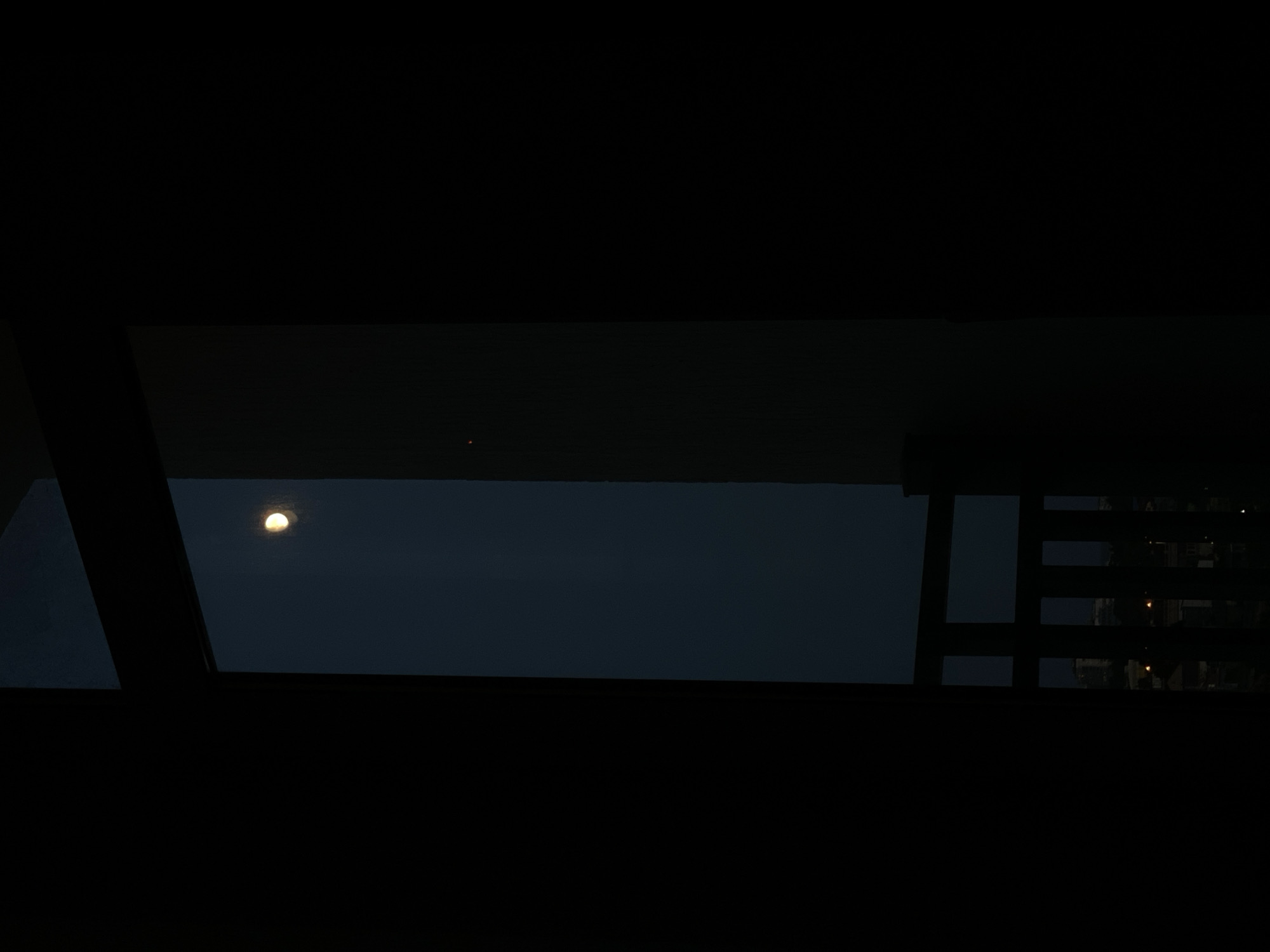 The image depicts a dark, nighttime scene viewed through a window. A bright, partially illuminated moon is visible in the upper central portion of the frame, set against a dark blue sky. To the right of the moon, a faint, reddish-orange light can be seen. The foreground is dominated by dark, silhouetted shapes, likely curtains or window frames, that obscure much of the view. In the lower part of the image, through the window's vertical bars, faint lights of a city can be discerned, suggesting an urban environment. The overall mood is somber and quiet, with the moon being the primary light source. The image does not contain any people or specific details that would allow for precise identification of the location within Sochi, Russia.