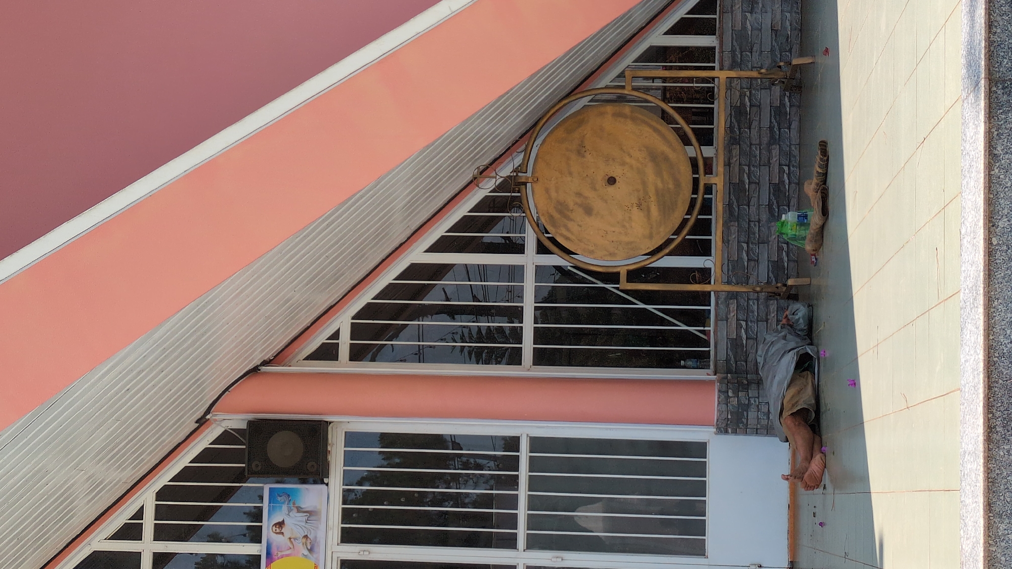 The image depicts the exterior of a building in Đa Thọ, Vietnam. The building has a distinctive architectural design with a sloping roof section and a large, round, ornamental gong mounted on the exterior wall. A man is lying on the ground next to the building, covered by a gray cloth. A pair of boots are situated near his head, along with a plastic bag. The ground is tiled, with some purple items scattered nearby. The building's façade consists of a combination of windows with horizontal bars, a speaker, and a poster.