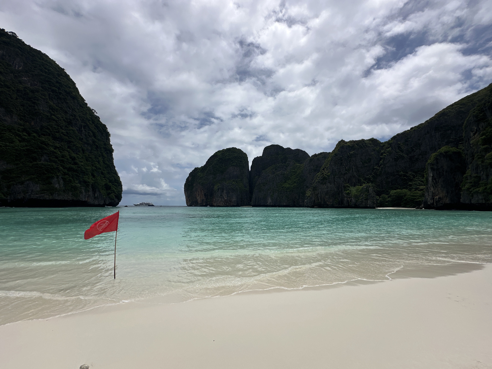 This image depicts a tropical beach scene, likely in Ao Nang, Thailand, characterized by its pristine white sand, clear turquoise waters, and dramatic limestone cliffs covered in lush vegetation. A red flag with a white symbol, possibly indicating a warning or prohibition, is planted in the shallow water near the shore. In the distance, a large white boat is visible on the calm sea, and several towering, verdant cliffs form a natural amphitheater around the bay. The sky is filled with a dramatic display of clouds, suggesting an overcast or partly cloudy day, but the overall atmosphere is serene and picturesque. The gentle waves lapping at the shore create a sense of tranquility.