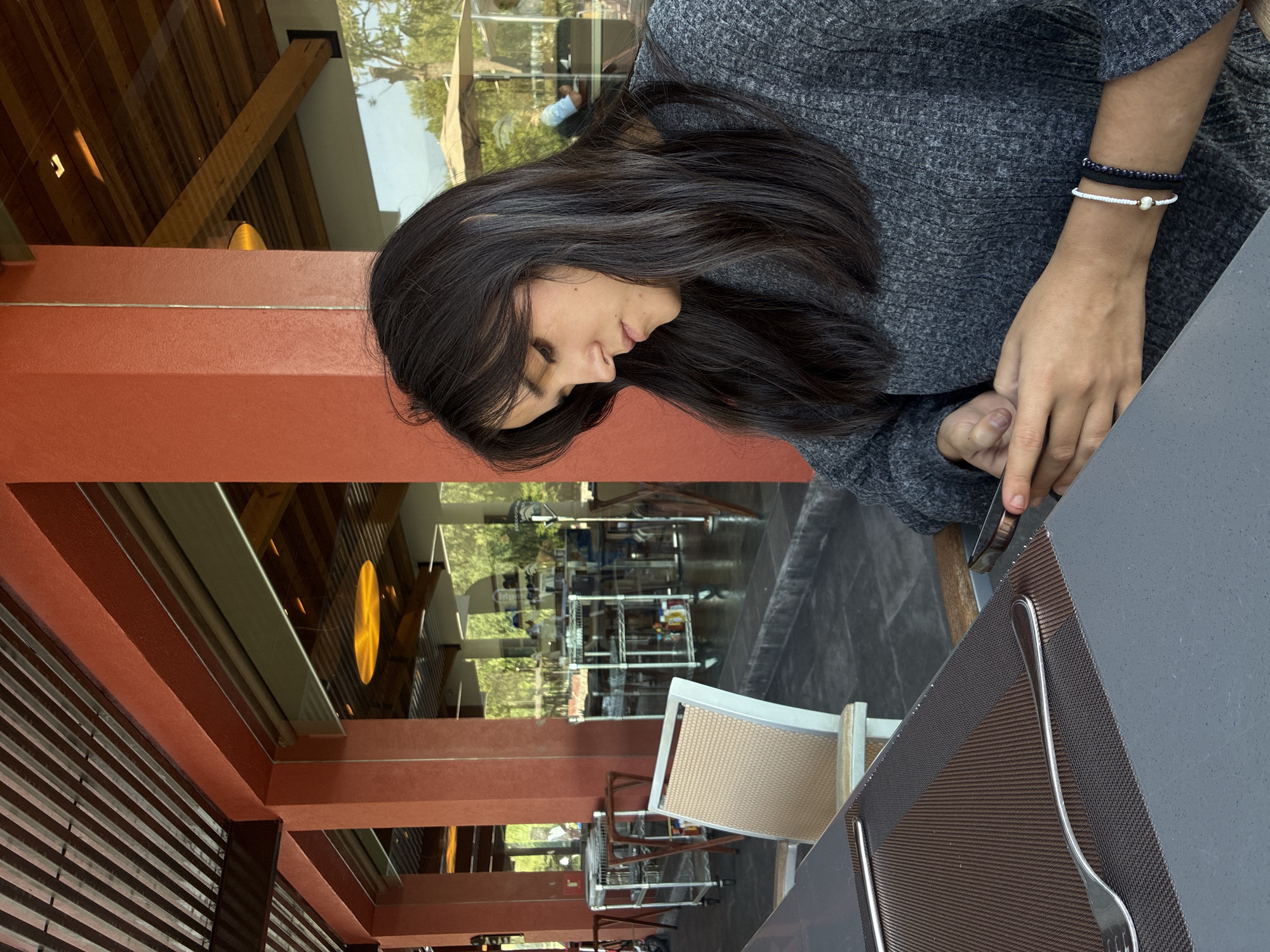A woman with long, dark hair wearing a grey sweater is seated at a table, appearing to be using a cell phone. She has two bracelets on her left wrist. The table has a dark grey surface and a brown woven placemat with silverware. The setting appears to be an open-air restaurant with a view to the outside, possibly in Pueblo Nuevo de Jasso, Mexico, given the request. The restaurant has a structure with red-brown pillars and wooden beams. Visible through the restaurant are trees and possibly other buildings.