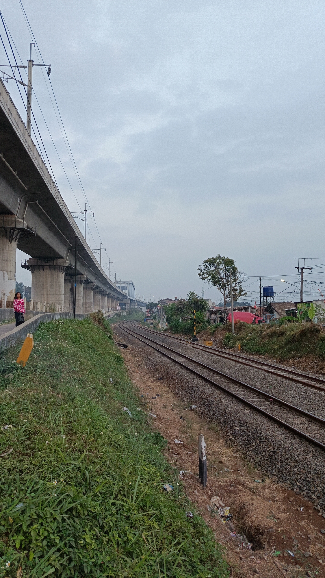 A woman in a pink and black patterned jacket stands on a concrete ledge next to a grassy slope. The ledge is part of an elevated train track structure, supported by numerous concrete pillars that extend into the distance. A yellow marker is positioned on the ledge to the left of the woman. The train tracks run parallel to the ledge, curving into the distance. Beyond the tracks, a densely populated residential area is visible, with houses of various sizes and colors, some with corrugated roofs, interspersed with trees and vegetation. A large blue water tank sits atop one of the buildings. Overhead power lines crisscross the scene. The sky is overcast with light grey clouds. The overall impression is an urban environment with infrastructure for transportation juxtaposed with informal settlements. The location is specified as Margajaya, Indonesia.