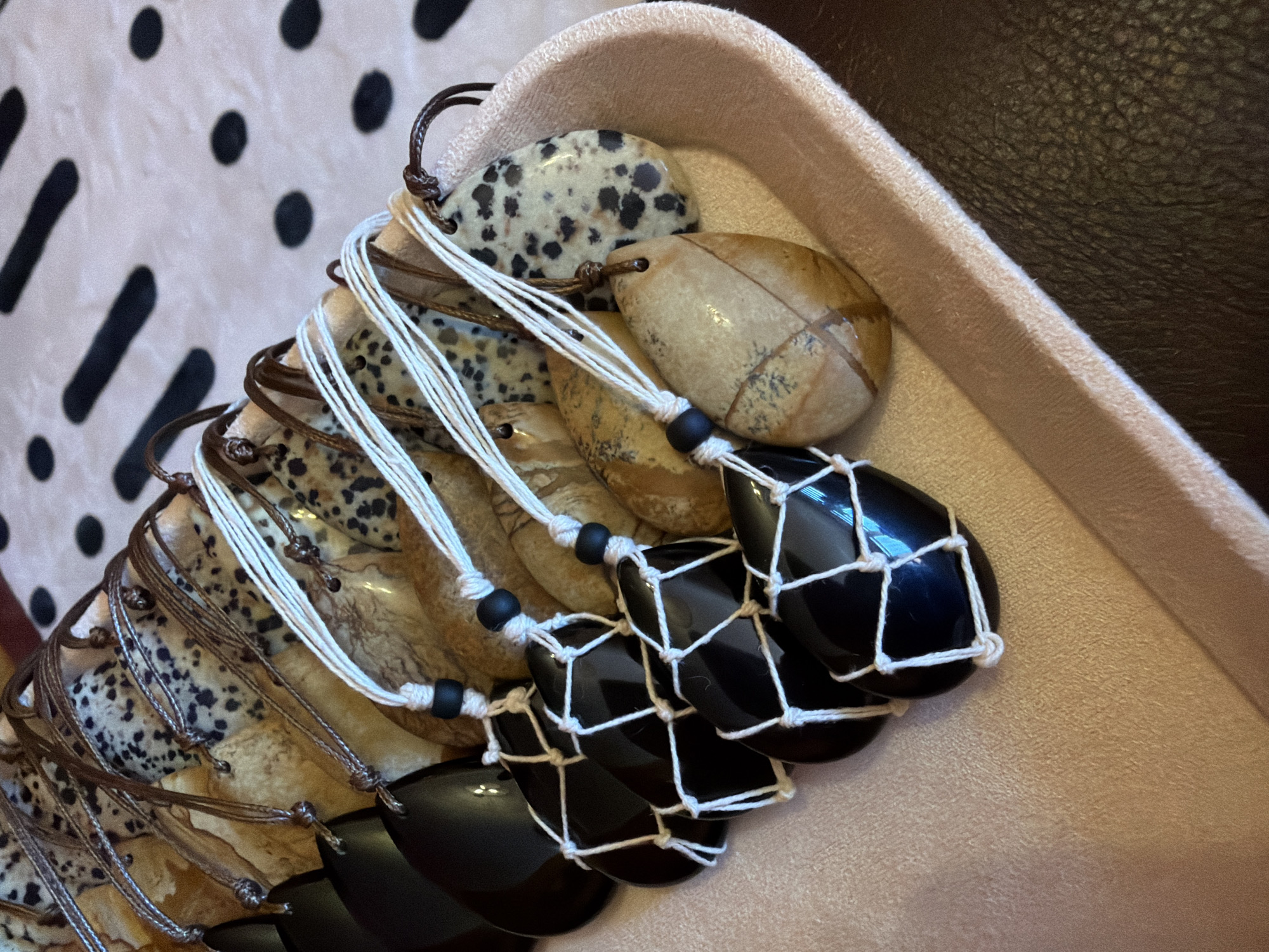 A shallow, light pink velvet-lined tray contains a collection of polished, teardrop-shaped stones with drilled holes and attached cords, arranged in a diagonal line from front left to back right. Some stones are a glossy black, while others are a mottled tan and brown, resembling picture jasper. The black stones are partially encased in white macrame netting, with a single black bead above each. The tan and brown stones are either uncased or have brown leather cords threaded through their holes. In the background, a fabric with a pattern of black dots and elongated black dashes on a white base is visible. The lighting suggests an indoor setting, likely a retail display or personal collection. There are no people or actions depicted. The location in Khimki, Russia is not discernible from the image.