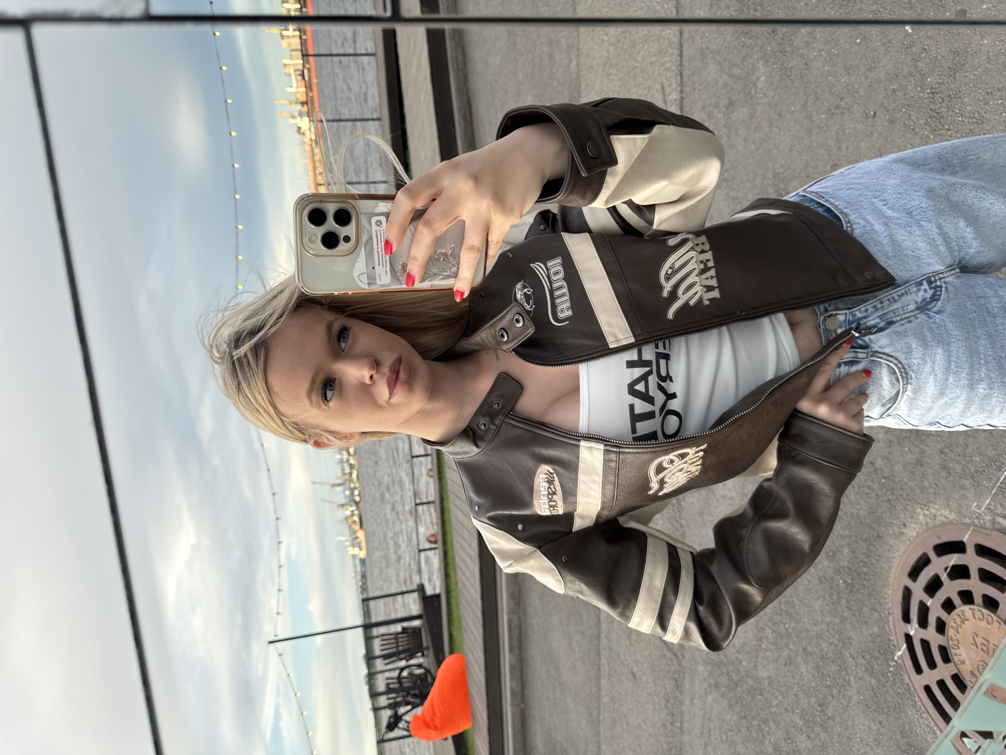 The image shows a young woman taking a mirror selfie outdoors. She is wearing a brown and cream-colored faux leather jacket with racing stripes and patches, a white tank top, and light-wash jeans. Her hair is blonde and she has red nail polish. The background is a slightly overcast sky reflecting in a mirrored surface, with a hint of a cityscape and a body of water visible. String lights are visible in the background, suggesting it might be evening or early morning. The setting appears to be a waterfront promenade or outdoor seating area. The overall mood is casual and confident. Visible text on her tank top reads "BETA BOYS". Text on a patch on her jacket is difficult to read, but appears to be a logo or brand name.