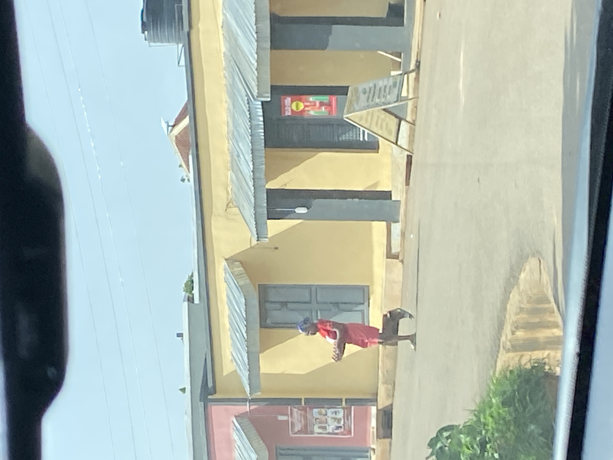 The image depicts a street scene in Bar Jirgi Summa, Nigeria. A woman wearing a red dress and a blue headscarf is walking across the paved road, carrying a tray of items. She is positioned in front of a yellow building with a corrugated metal awning. To the left of the yellow building is a pink building with a similar awning. There are two A-frame signs on the pavement in front of the yellow building. The sky is clear and blue, and power lines are visible stretching across it. Green foliage is present in the lower left corner of the frame.