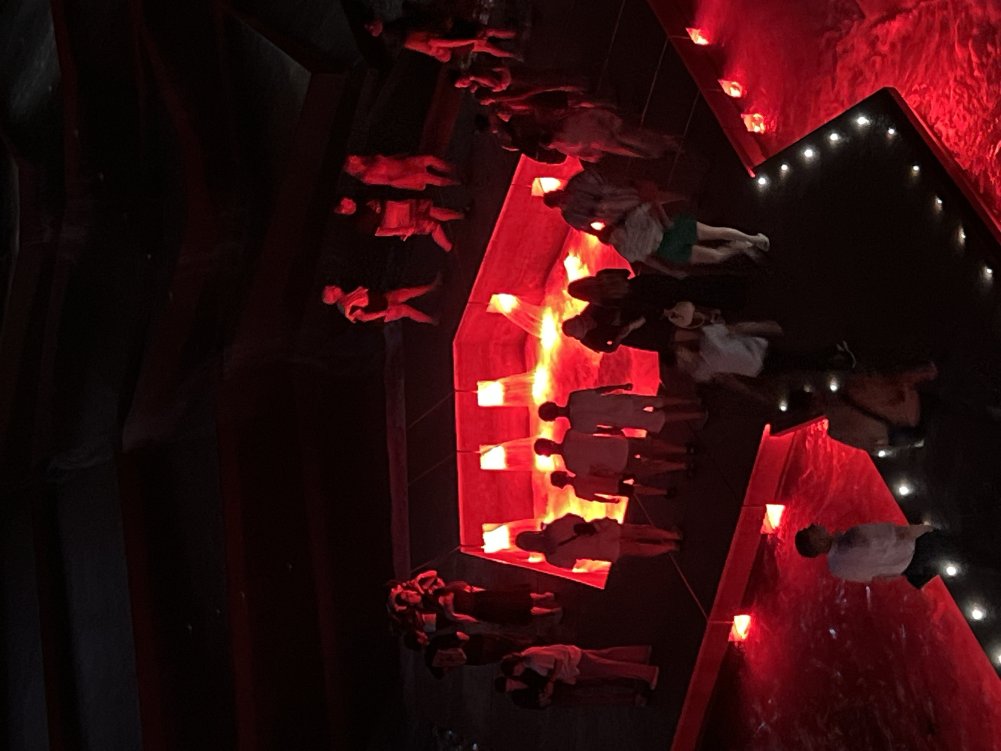 The image depicts an aerial view of an art installation or event, predominantly illuminated by intense red light. Several groups of people are present, standing and walking on dark platforms surrounding a central, rectangular pool filled with red liquid or light. The pool has bright, square light sources embedded within its structure. The surrounding environment appears to be a dark, enclosed space, possibly an exhibition hall or outdoor venue at night. The people are dressed in casual clothing, and their poses suggest they are observing the installation or interacting with each other. The overall atmosphere is dramatic and somewhat mysterious due to the low lighting and the striking red hues. There is no specific indication that this scene is located in the city of Krasnodar, Russia; the details provided are purely based on the visual information within the image itself.