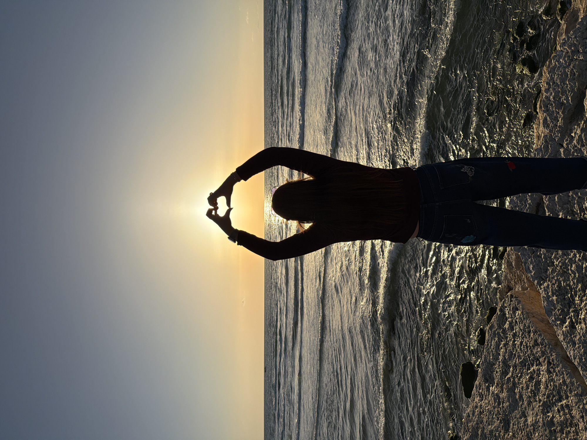 A vertical outdoor shot shows a female figure silhouetted against the sun at what appears to be a beach at sunset. The scene is possibly in Tel Aviv, Israel, given the presence of a beach. The woman is wearing dark jeans and a dark long-sleeved shirt. She is standing with her arms extended overhead, her hands forming a heart shape around the sun. The horizon line is visible behind her, with the sky to the left, transitioning from light to dark, and the sea to the right. The waves are visible, and they are reflecting the light. Rocks or cliffs are visible in the lower right corner.