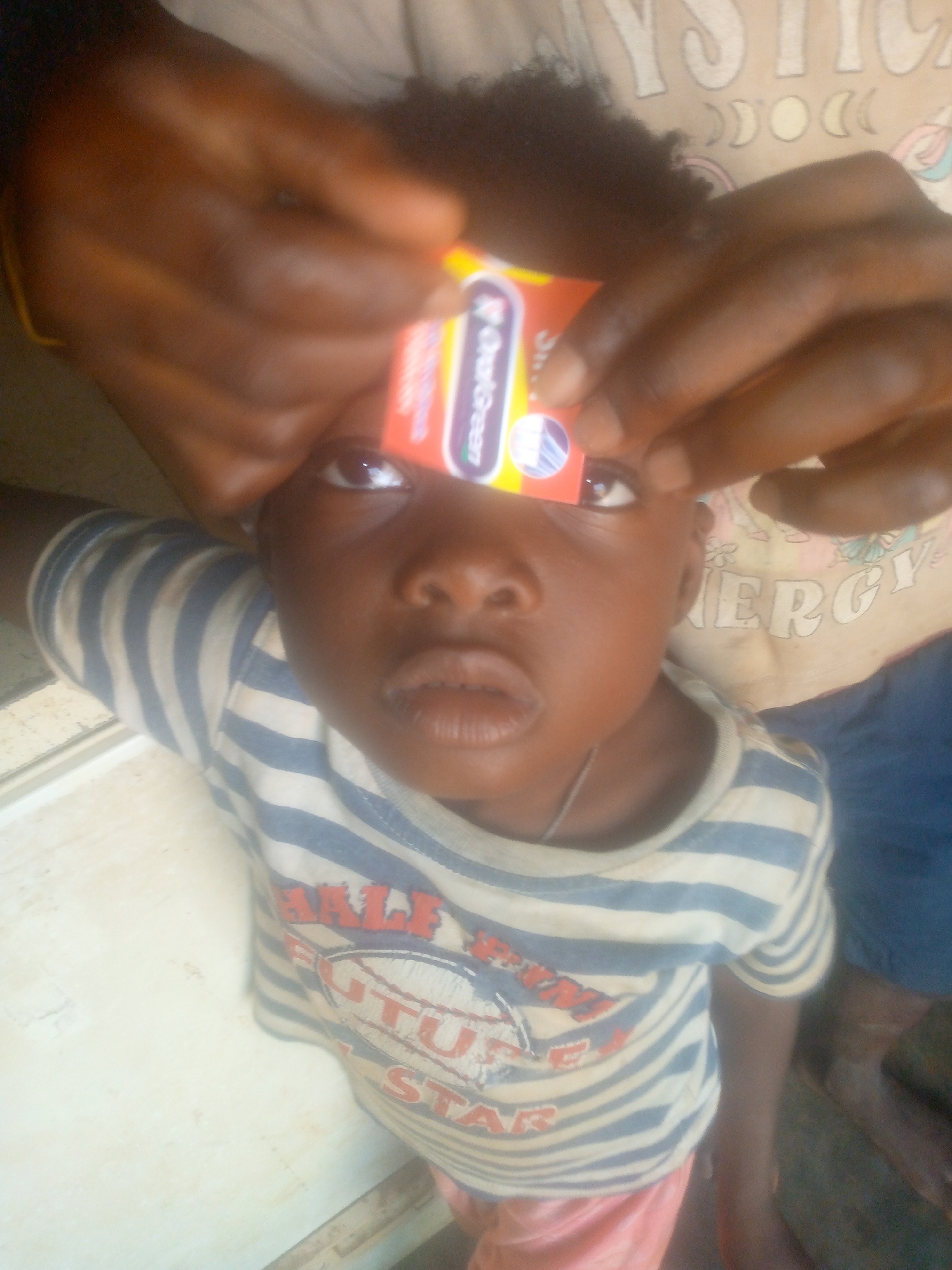 A young child with dark skin and short, dark, curly hair looks directly towards the viewer in a close-up, high-angle shot. Above the child's head, an adult's dark-skinned hands are visible, holding a small, rectangular, brightly colored package in front of the child's eyes, partially obscuring the upper part of their face. The child's eyes are looking upwards past the object, and their lips are slightly parted.
The child is wearing a light-colored t-shirt with horizontal blue stripes. On the front of the shirt, red and white text and graphics are visible, including partial words like "FUTURE" and "STAR." They are also wearing pink shorts. In the background, part of an adult's light-colored t-shirt is visible, displaying white text that includes "MYSTIC" (with associated crescent moon and circle symbols) and "ENERGY."
The package held by the adult's hands is predominantly red, orange, and yellow. Clearly visible text on the package, set against a green background, reads "EverGreen." The background of the scene is indistinct, suggesting an indoor or sheltered setting, with a light surface to the left and a darker area to the right. The child's expression appears neutral or slightly curious.