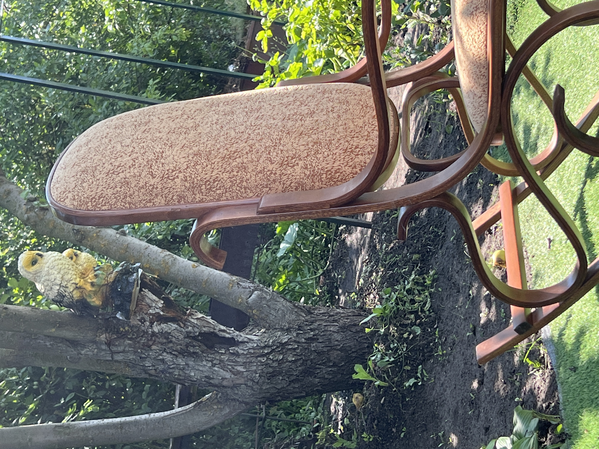 In Khryashchevka, Russia, an ornate wooden rocking chair with a patterned upholstered seat and back is positioned on artificial turf in a garden setting. Behind the chair, a tree trunk with rough bark is visible, from which a decorative statue of an owl perched on a small base emerges. The background consists of lush green foliage and a dark garden bed.