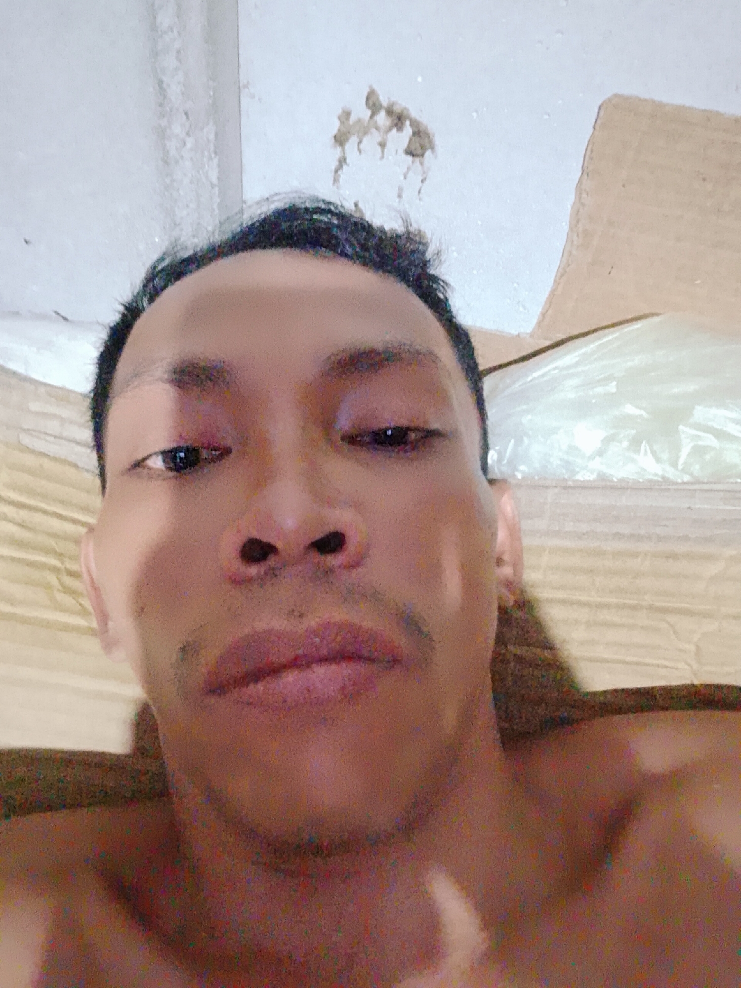 A close-up selfie of a dark-skinned man with short black hair. He has dark eyes, a flat nose, and thin lips. A faint mustache is visible. The background includes a white ceiling with some discoloration or debris near the top, along with what appears to be cardboard and plastic packaging. It is not possible to confirm the location as Bukit Ketok, Indonesia from this image alone.