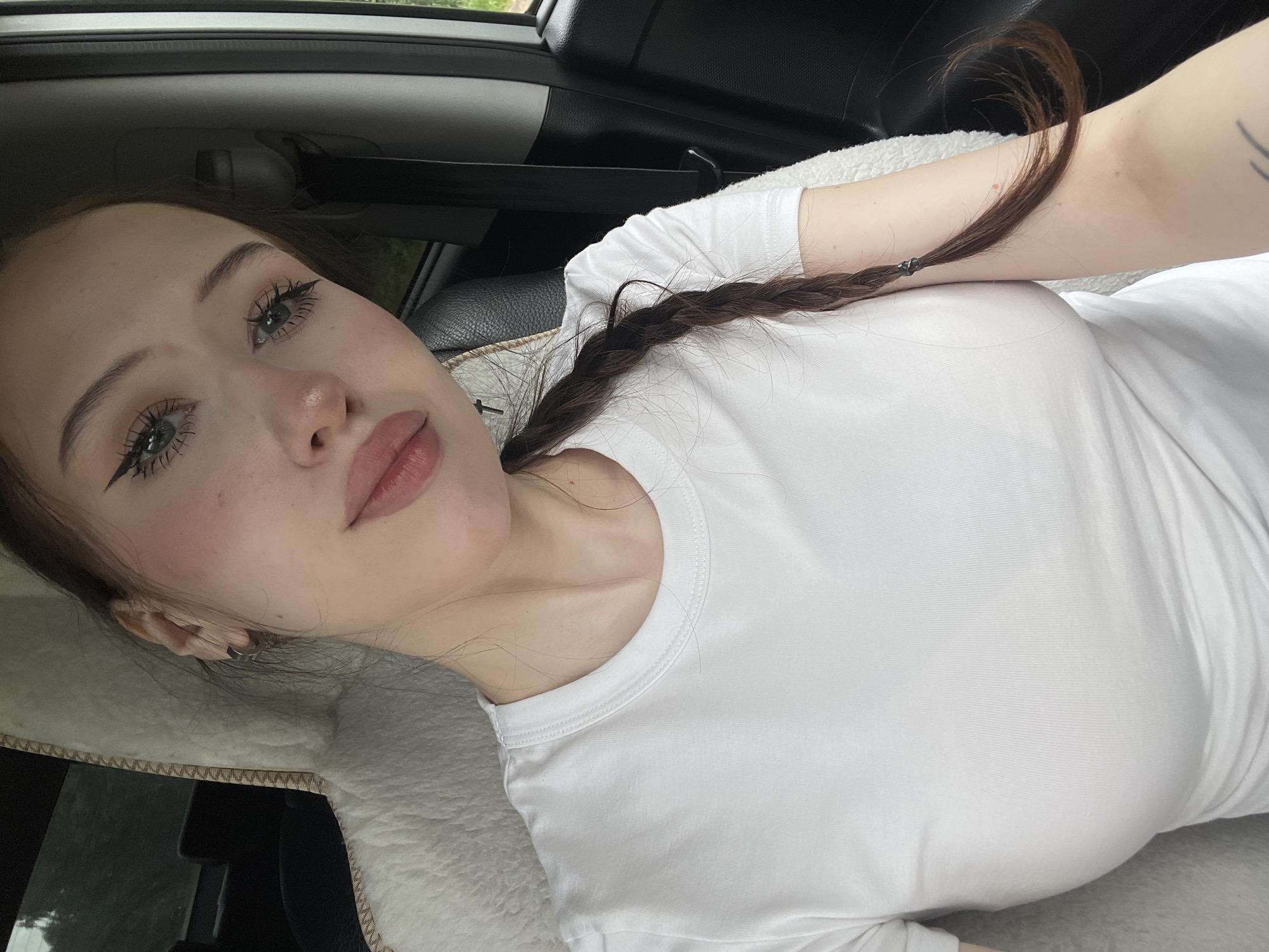 A young woman with fair skin, dark hair braided to her right, and light brown eyes is pictured inside a vehicle. She is wearing a white t-shirt. The woman has winged eyeliner and pink blush on her cheeks. Her hair is tied with a dark elastic band. The background shows the interior of a car, including a fuzzy white seat cover on the left and black leather on the right, with a black seatbelt visible. The scene is outdoors, as indicated by the greenery visible through the car window on the right. The lighting suggests it is daytime. The image was taken in Gavrilovka, Russia.