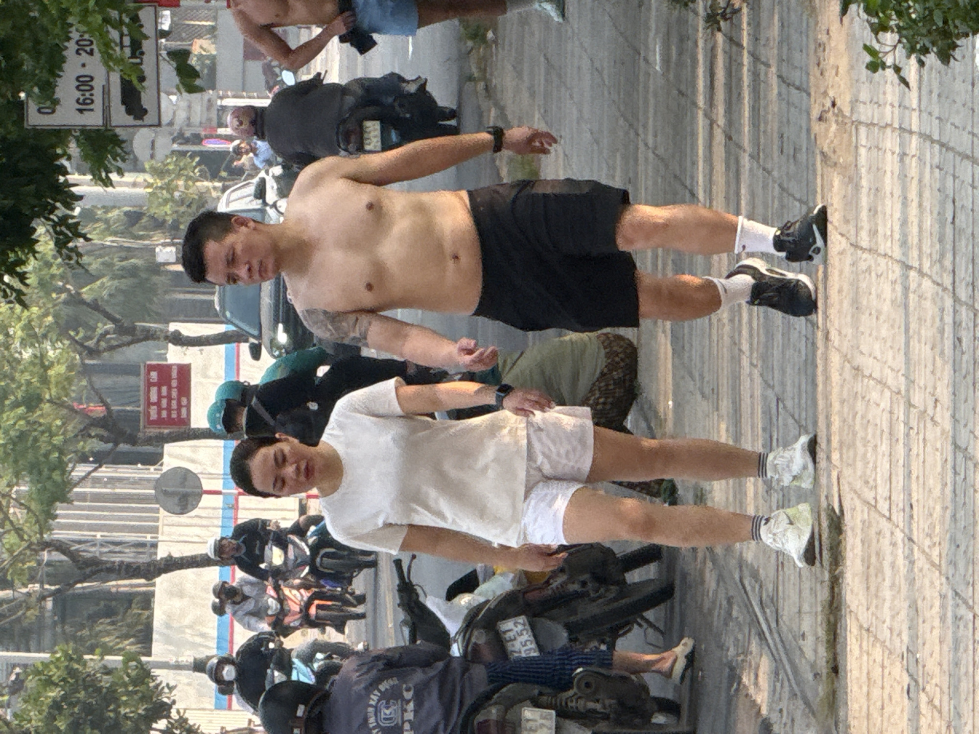 On a paved sidewalk in what appears to be Đà Nẵng, Vietnam, stands a shirtless man in black shorts, black athletic shoes, and white socks. Next to him is another man in a white t-shirt, white shorts, white socks, and light-colored athletic shoes. Both men wear wristwatches on their left wrists. In the background, there are motorbikes parked along the curb. People wearing helmets are on some of the motorbikes. A sign restricting bus traffic is visible in the upper left corner of the image. Trees and buildings line the street.