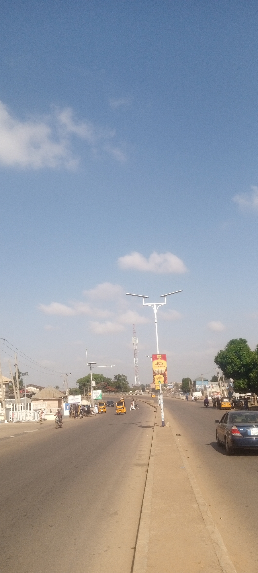 The image shows a street scene in Sabon Tasha, Nigeria on a sunny day with a clear blue sky with scattered clouds. The foreground shows a wide, paved road with a raised sidewalk on the right. Cars, motorcycles, and auto-rickshaws ("keke napeps") are seen on the road. Several pedestrians are present. A tall, white, modern lamppost with two horizontal lights is prominent along the sidewalk. A banner with a food advertisement is attached to the lamppost. A cellular communication tower is visible in the background, behind the lamppost. Buildings line the left side of the road, partially obscured by trees.