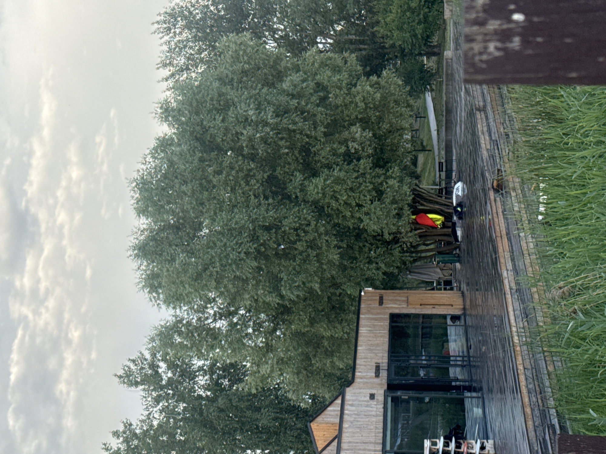 The image captures a waterside scene, possibly in Nizhny Novgorod, Russia, with a modern wooden building featuring large glass windows. The building appears to be a restaurant or cafe, with seating visible inside. In front of the building is a wooden deck, which extends towards the viewer and is partially covered by lush green grass on the right. On the deck, near the building, there are some kayaks or canoes, one with a red and yellow cover, suggesting recreational water activities. A wooden railing runs along the right edge of the deck. The background is dominated by large, leafy green trees, and the sky is overcast with soft, diffused light, indicating a cloudy day, possibly morning or late afternoon. The overall atmosphere is peaceful and serene, with a focus on nature and modern architecture. No visible text is present in the image.