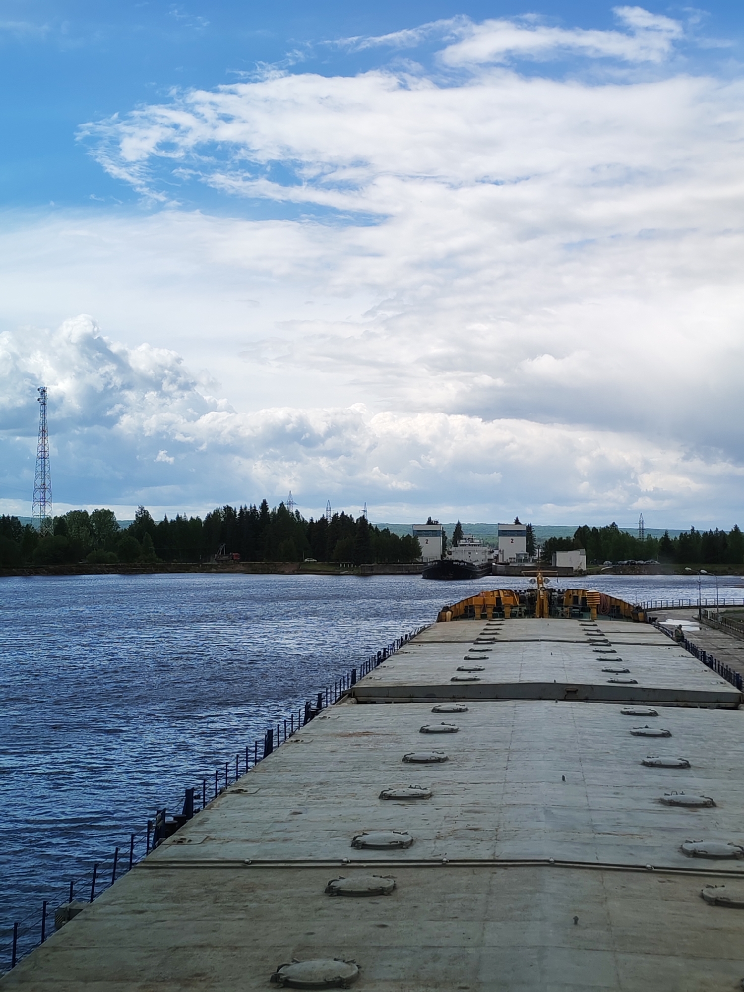 The image shows a view from a barge or large boat, likely in Белоусово, Russia. The barge has a flat, concrete deck with round hatches and is bordered by a railing. The water is a dark blue. Across the water is a green, forested shoreline with a tall communications tower to the left and what appears to be a building or structure with the number "2" on it. The sky is mostly cloudy, with patches of blue.