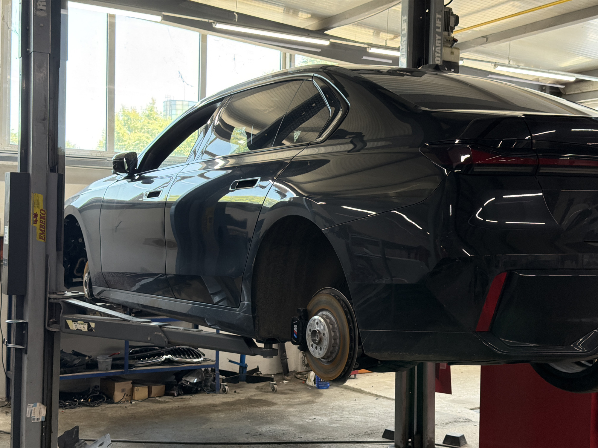 In Burgas, Bulgaria, a black sedan, identified as a BMW 7 Series (G70) judging by the distinctive taillight design and window trim, is elevated on a car lift in a well-lit garage. The vehicle's rear wheel on the driver's side is removed, revealing the brake assembly, which features a caliper with an "M" logo. The car is positioned within a workshop environment, indicated by the presence of tools, shelving units holding car parts, and an overhead lighting system. The overall scene suggests a car maintenance or repair operation.