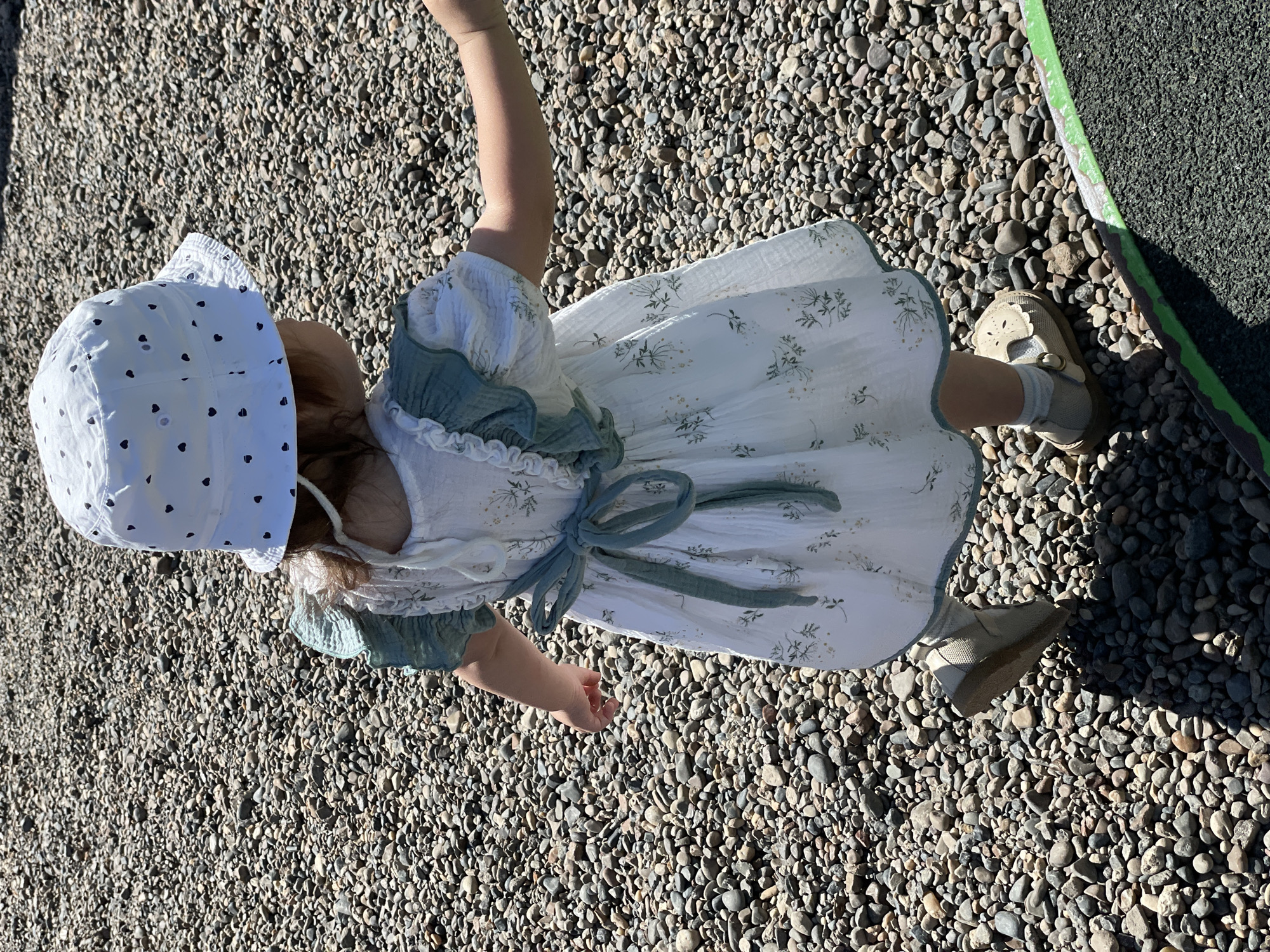 A toddler is standing on a bed of small, gray pebbles. The toddler is wearing a white dress with a floral pattern and a light green trim around the neckline and sleeves, and it is tied with a light green bow. She is also wearing a white bonnet with small black hearts. The toddler is wearing tan shoes and white socks. The background includes a patch of green and black rubberized safety flooring on the right. The scene appears to be outdoors in daylight. There is no indication of the location being in Izhevsk, Russia.