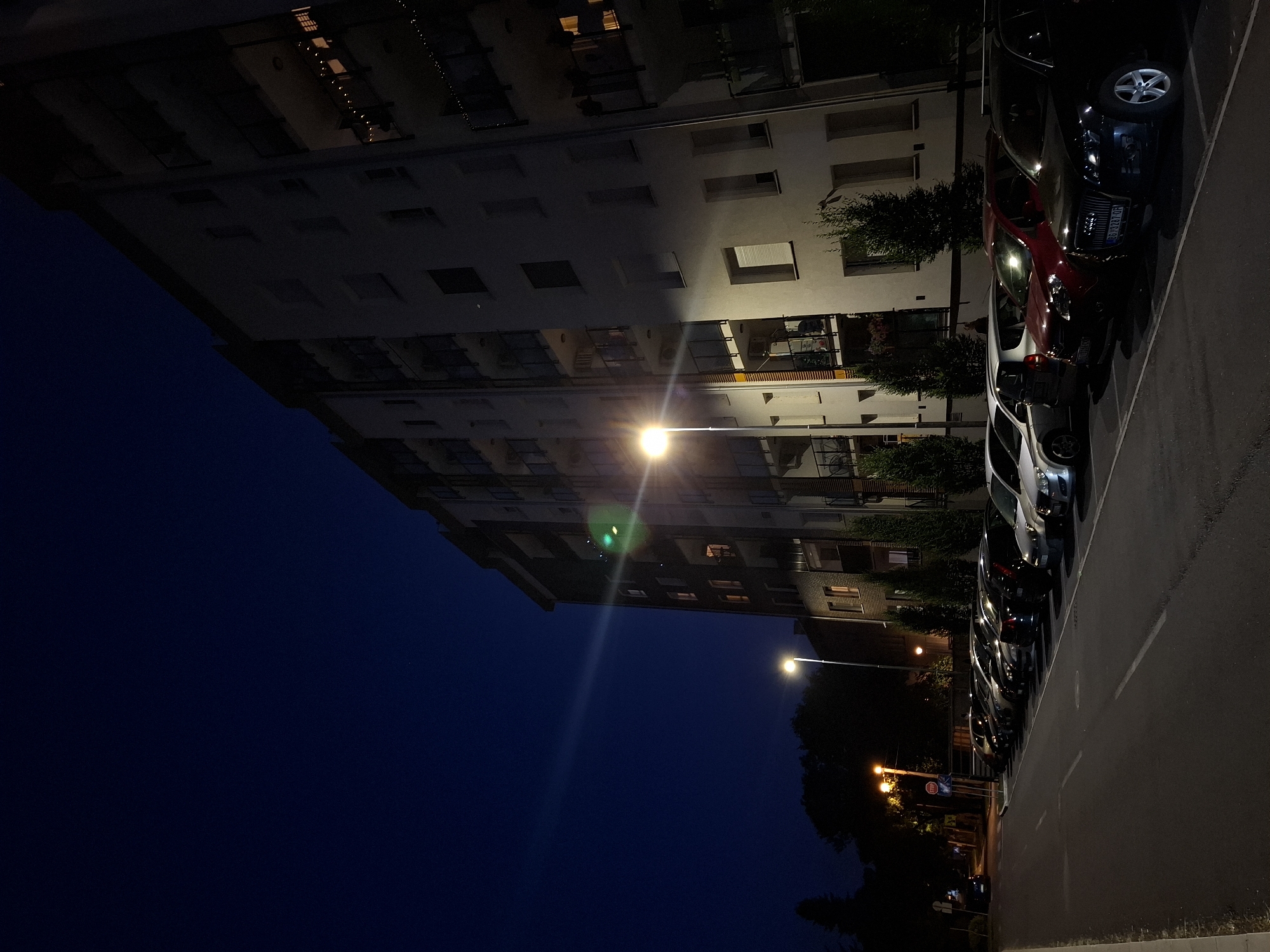 A nighttime view shows a modern apartment building on the right side of the frame, its facade illuminated by streetlights. The building has multiple balconies, some with string lights, and windows of various sizes. A street runs along the left side of the image, lined with parked cars. The sky is a deep, clear blue. Trees and a streetlamp are visible in the background. The scene appears to be a residential area in Zemun Urban Municipality, Serbia.
