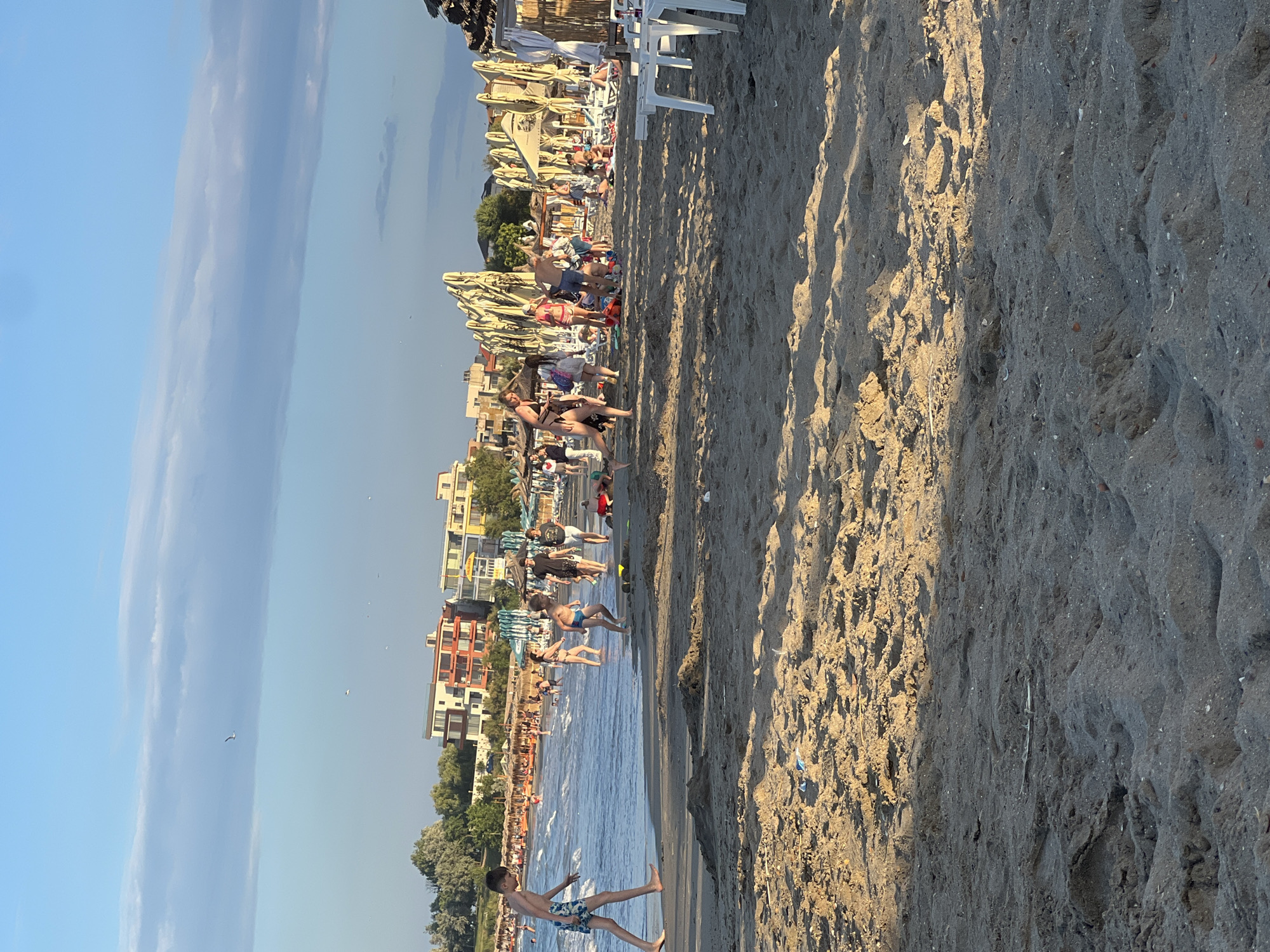 The image depicts a crowded beach scene on a sunny day in Constanța, Romania. In the foreground, the sandy beach is visible, with footprints and some scattered debris. Moving towards the background, people are spread across the sand and in the shallow water. Several individuals, including children, are engaged in various beach activities, such as walking, playing, and wading. Beach umbrellas and lounge chairs are set up in rows further back, indicating a resort or public beach area. In the distance, buildings, likely hotels or apartments, are visible along the coastline. The sky is a clear blue with some wispy clouds, and the sun casts long shadows on the sand. The overall atmosphere is lively and recreational.