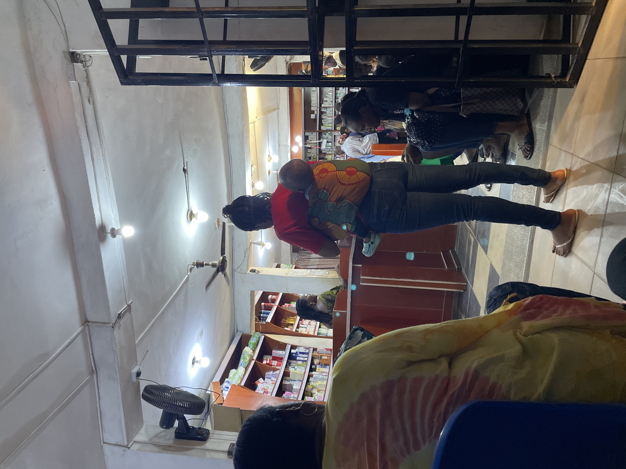 A pharmacy scene in Jos, Nigeria, with a woman carrying a child on her back. She is dressed in a red shirt and jeans, looking at medications on the shelves. The child is secured with a colorful African print carrier. The pharmacy has rows of shelves stocked with various medications and health products. Other customers are present, with one woman in a patterned dress standing in the foreground. There are ceiling fans and fluorescent lighting. A metal grid structure is visible near the ceiling, potentially for hanging displays. A black desk fan stands on the counter. There is a yellow and peach-colored fabric and a blue plastic chair. The flooring appears to be tiled.