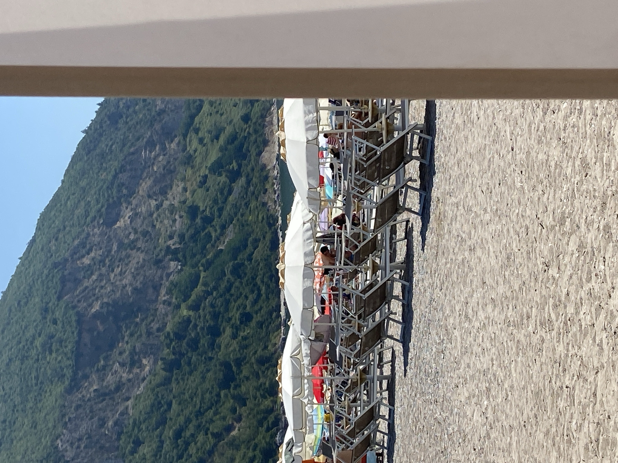 The image depicts a beach scene in Prijevor, Montenegro, with a focus on rows of lounge chairs and umbrellas set up on the sand. In the background, a lush green, rocky mountainside rises up, meeting a clear blue sky. Several white umbrellas with gold trim are prominently displayed, providing shade for empty lounge chairs. Some chairs are occupied by people, though they are not the primary focus. The beach itself is sandy, with subtle textures indicating ripples and slight unevenness. The perspective is from a slightly elevated angle, looking towards the mountain and the sea beyond. A portion of a light-colored structure, possibly a building or a cabana, partially obstructs the right side of the frame.