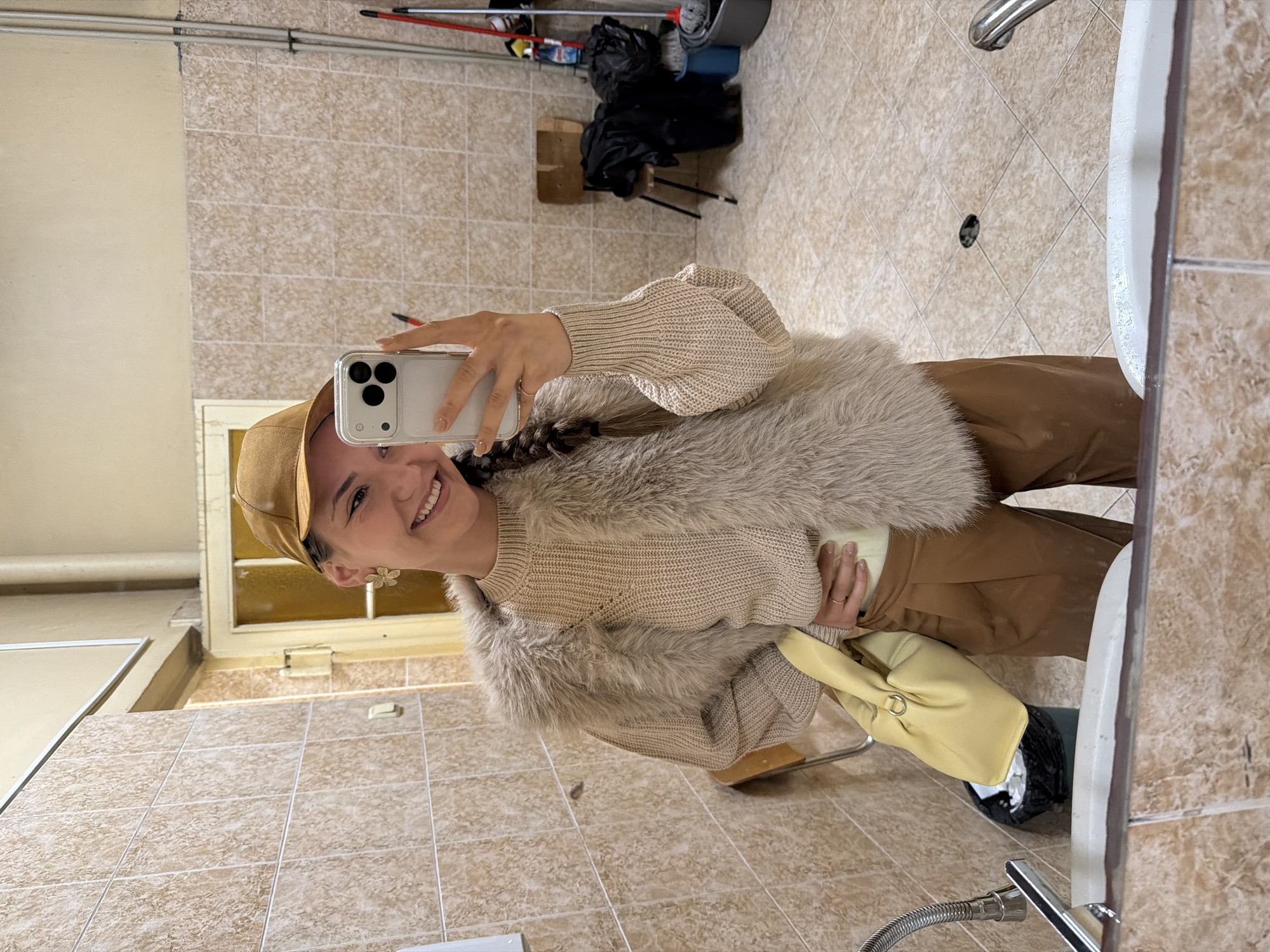 A woman is taking a selfie in a bathroom. She is wearing a tan baseball cap, a light tan sweater, a fur-like vest, and brown pants. The bathroom has beige tile walls and floor. In the background, there is cleaning equipment, including a mop and bucket. The presence of the equipment suggests that the bathroom is in a commercial or public space, but the location in Sofia, Bulgaria, cannot be determined from the image.