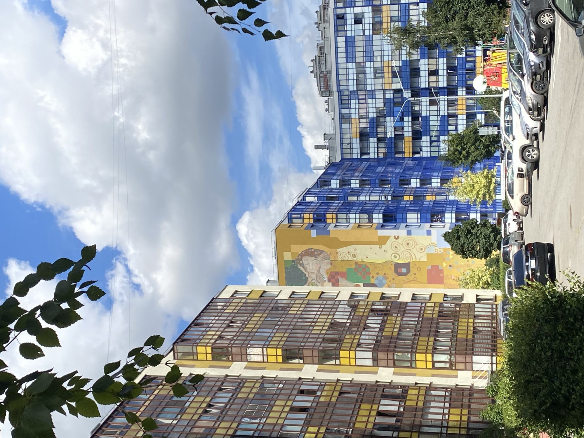The image depicts a view of a residential area in Kudrovo, Russia, under a partly cloudy sky. In the foreground, a dense green bush is visible on the left. Behind the bush, a black sedan is parked. Several other cars are parked in a lot, including silver SUVs and sedans, as well as a white van.
The middle ground is dominated by a row of multi-story apartment buildings. The building on the far left is light-colored with yellow and brown-tinted windows. Adjacent to it is a taller building with a large mural on its facade, featuring abstract shapes and figures in yellow, beige, and green. To the right of this is a brightly colored apartment building with blue and yellow window frames. Further to the right, another apartment building features a predominantly blue facade with yellow and white accents in its windows.
In the background, more apartment buildings of varying heights are visible. Trees are interspersed among the buildings, and streetlights are present in the parking lot. The sky is a clear blue with large, fluffy white clouds.