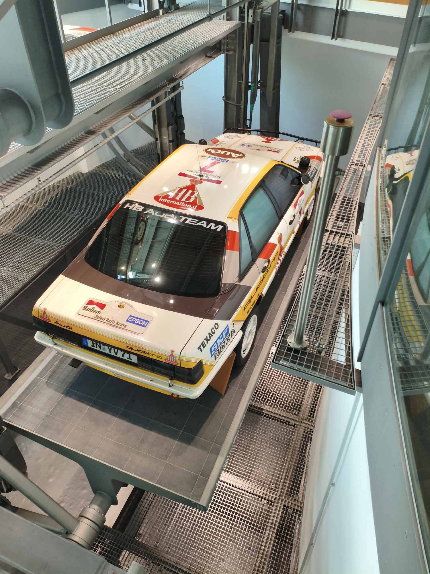 The image shows a white Audi Sport Quattro rally car, displayed on a raised platform within the Audi Forum Ingolstadt in Ingolstadt, Germany. The car, which has red and yellow racing stripes and various sponsor logos such as HB, International, Marlboro, Safari Rally Kenya, and Texaco, is positioned on a grey tiled surface with a metal grate walkway visible to the right and left of the platform. A cylindrical metal support structure with a pink cap is located to the right of the car. The background consists of a light blue wall and metal grating structures. There are no people visible in the image.