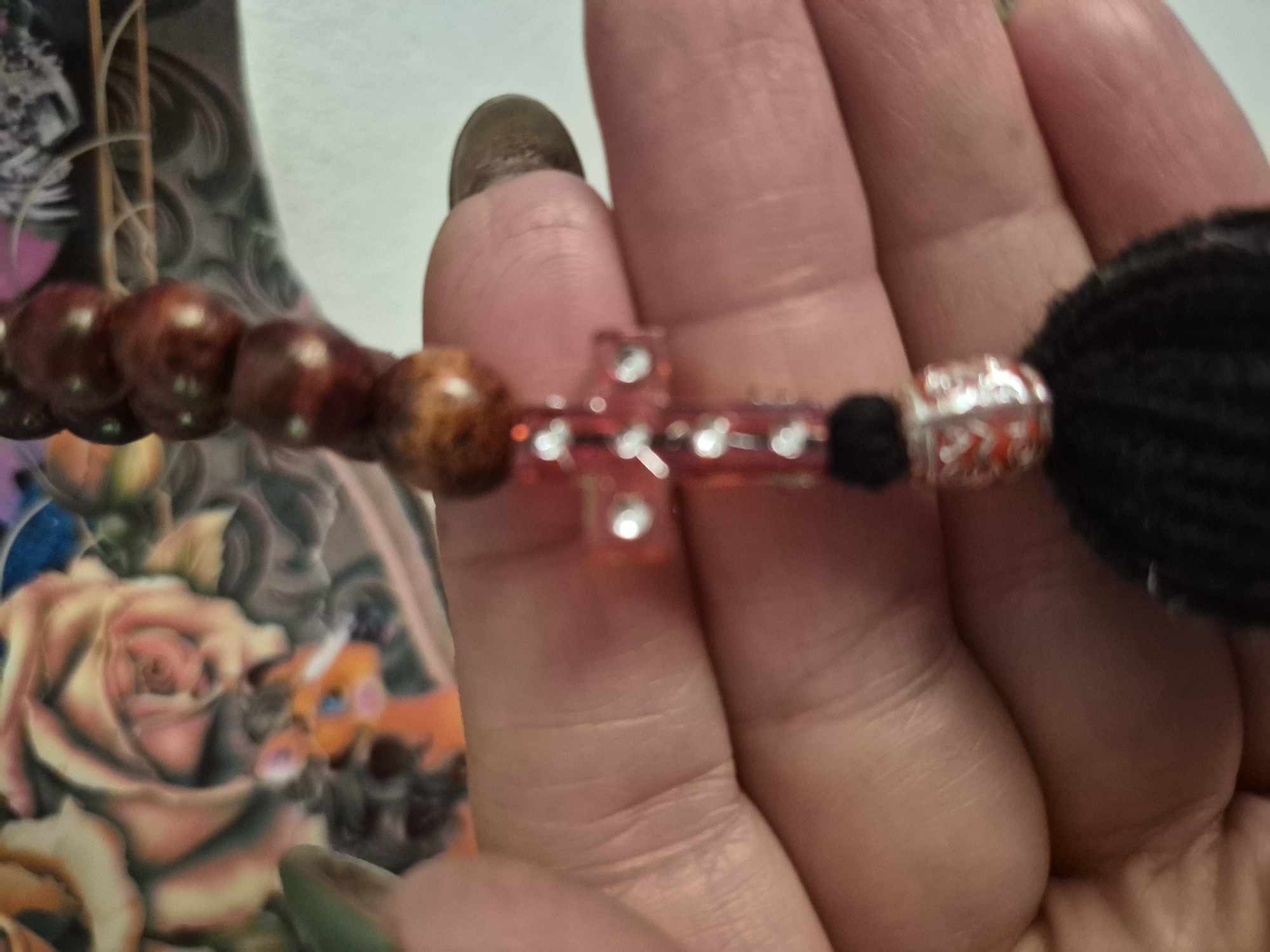The image shows a close-up of a person's hand holding a string of prayer beads. The beads are reddish-brown and are separated by a small, red cross adorned with clear stones. At the bottom of the string is a black tassel with a silver and red decorative bead. In the background, a blurred image of a tattoo depicting roses and a small animal can be seen. The image is taken from a high angle, and the lighting is natural. The person's skin is visible, with some nail polish on their fingernails. The setting is unclear but appears to be indoors. There is no indication of the location being in Vanadzor, Armenia.