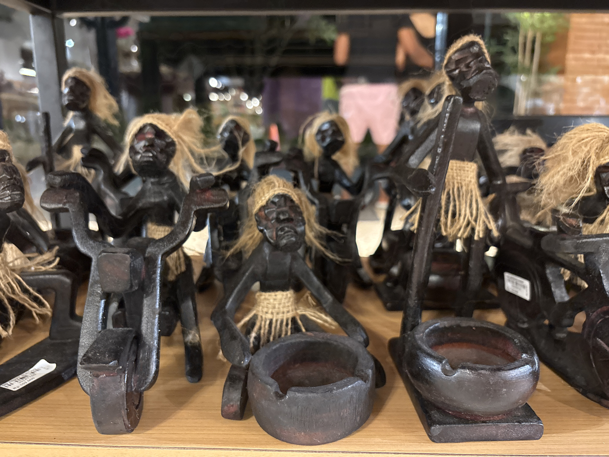 The image shows a display of dark-colored, carved wooden figures on a light-colored wooden shelf. The figures appear to be depictions of people, each with raffia-like hair and a raffia skirt. Some of the figures are situated with miniature ashtrays, while others are positioned with small, primitive-looking wheeled vehicles. The backdrop is blurred, suggesting a retail environment with other merchandise and a display window. The location is Kuta Selatan, Indonesia.