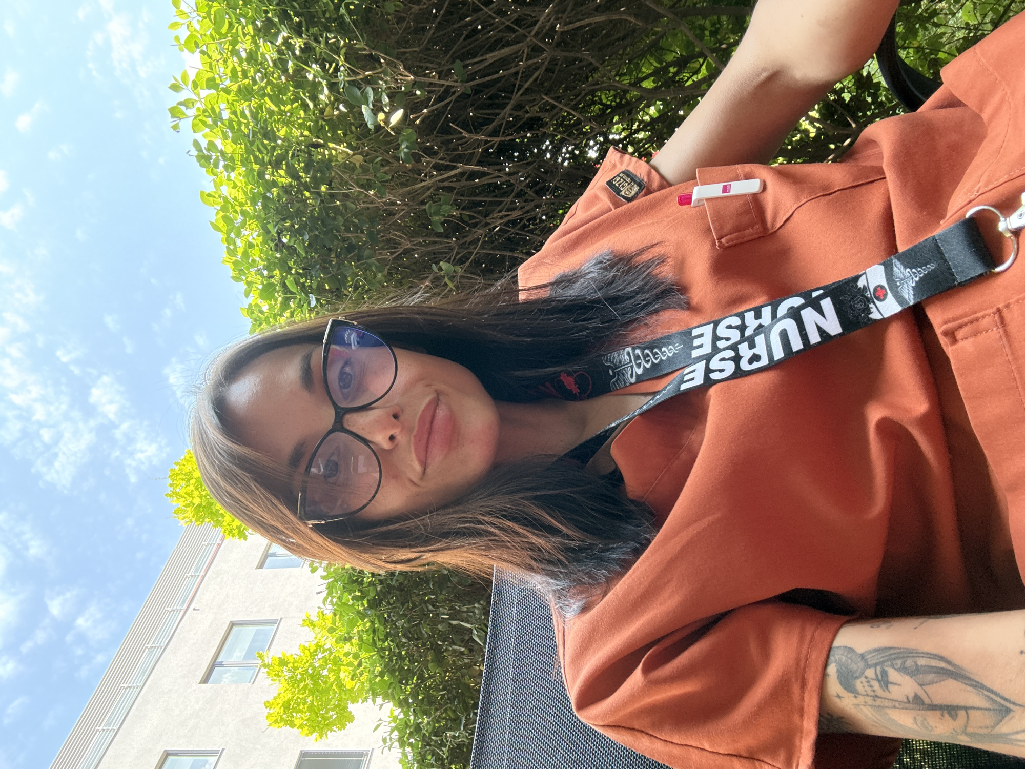 A woman with dark hair and glasses is taking a selfie outdoors, sitting in a chair. She is wearing an orange scrub top with a lanyard around her neck that says "NURSE". A pen is visible in her scrub top pocket. Behind her, there are green bushes, a building, and a blue sky with clouds. The location is Pleven, Bulgaria.