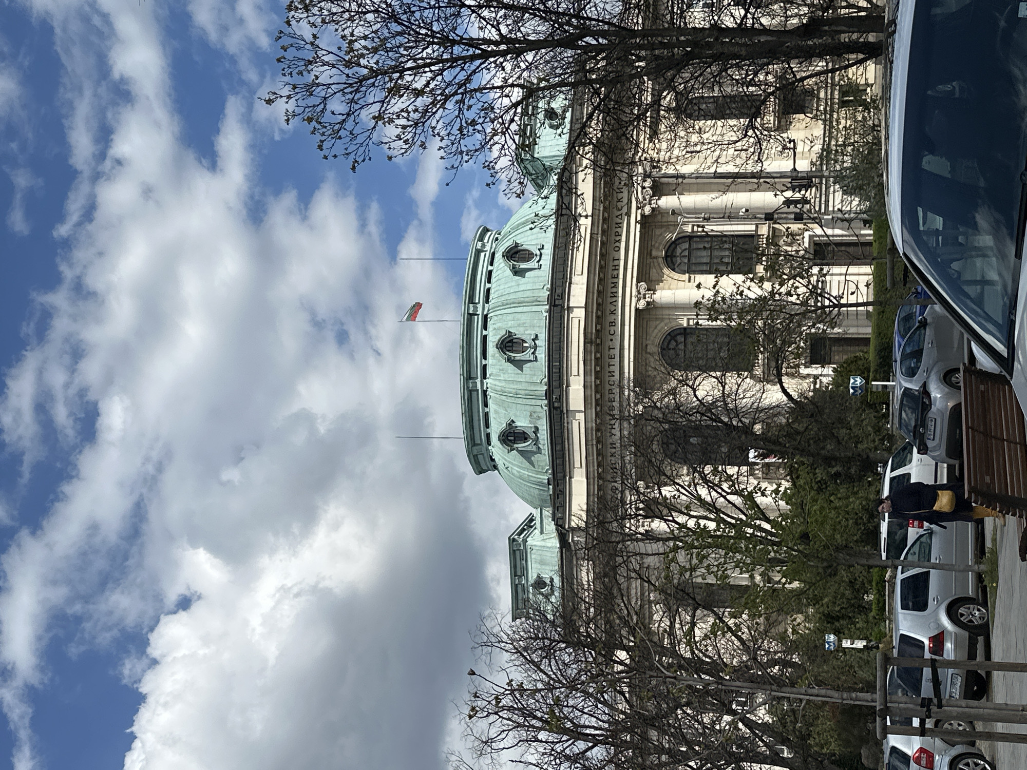 The image shows a building with a green dome, likely a landmark in Sofia, Bulgaria, based on the presence of the Bulgarian flag. The building's facade is white and features architectural details such as pillars and arched windows. The text "УНИВЕРСИТЕТ.СВ.КЛИМЕНТ.ОХРИДСКИ" is visible on the building, suggesting it is associated with the University of Sofia. Bare tree branches partially obscure the building and sky. The sky is partly cloudy with a mix of blue sky and white clouds. Cars are parked along the side of the road, and a person wearing a dark coat and yellow bag is walking near the cars. A portion of another vehicle is visible on the right side of the image.