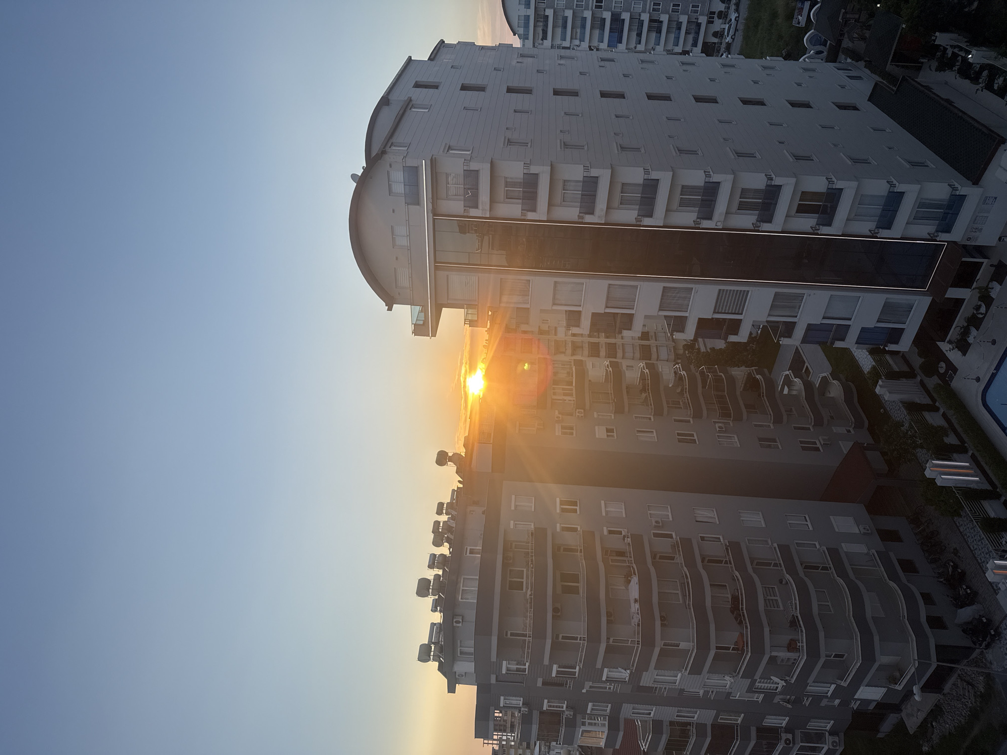 The image displays a modern urban landscape at either sunrise or sunset, with the sun low on the horizon, casting a bright golden light and lens flares. The scene is dominated by several multi-story buildings, likely residential or hotel complexes, under a clear sky that transitions from a soft blue to a warm yellow-orange around the sun.
In the mid-ground, a prominent tall building with a light grey facade features numerous balconies and a distinctive rounded roofline at its peak. A dark, reflective, vertical strip is visible on its side. Sunlight strongly illuminates one side of this building, while the other side is in shadow. Behind it, another similar building is partially visible.
To the left of the main structure, another multi-story building with a light-colored facade and wavy-fronted balconies is seen. Its rooftop is lined with multiple cylindrical objects, which appear to be solar water heaters, glinting in the direct sunlight. Shadows deepen towards the bottom and right of the frame, revealing glimpses of a swimming pool with blue tiles, some trees, and paved areas, suggesting an organized complex. The time of day is clearly the golden hour, characterized by the intense, warm light. No individuals or discernible text are visible in the image.