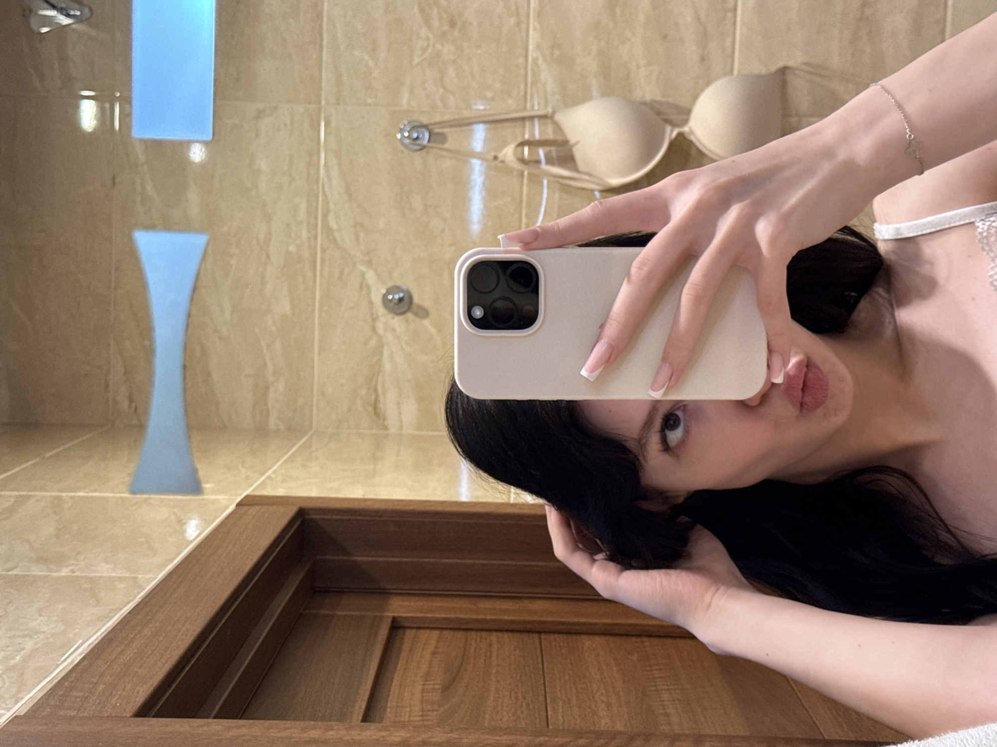 The image shows a young woman with black hair taking a selfie in a bathroom. She is holding a light-colored smartphone in her right hand, with her left hand touching her hair. She is making a kissing face. The background shows beige tiled walls, a wooden door frame, and a bra hanging on a hook. The setting appears to be a private residence. There is no information in the image to suggest the location is in the city of Khimki, Russia.