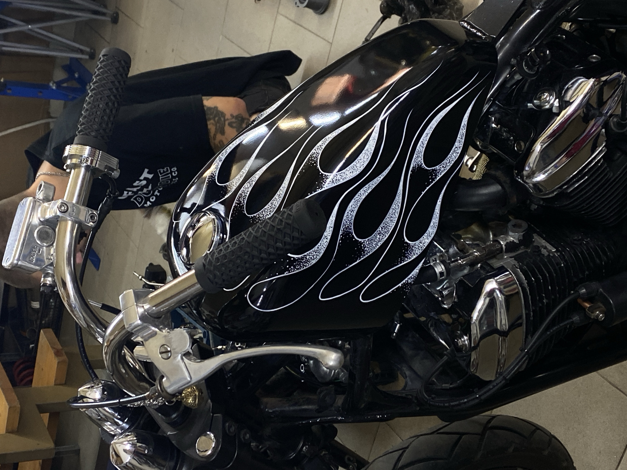 The image shows a close-up of a black motorcycle with a flame design on its gas tank. The motorcycle is parked indoors, possibly in a workshop or garage, given the presence of tools and equipment in the background. A person, partially visible from the back, is wearing a black t-shirt and appears to be working on the motorcycle. The setting is industrial, with concrete floors and metal shelving. The overall impression is one of a customized or vintage motorcycle being maintained or modified. No specific details about the location in the city of Agronom, Russia are discernible from the image.