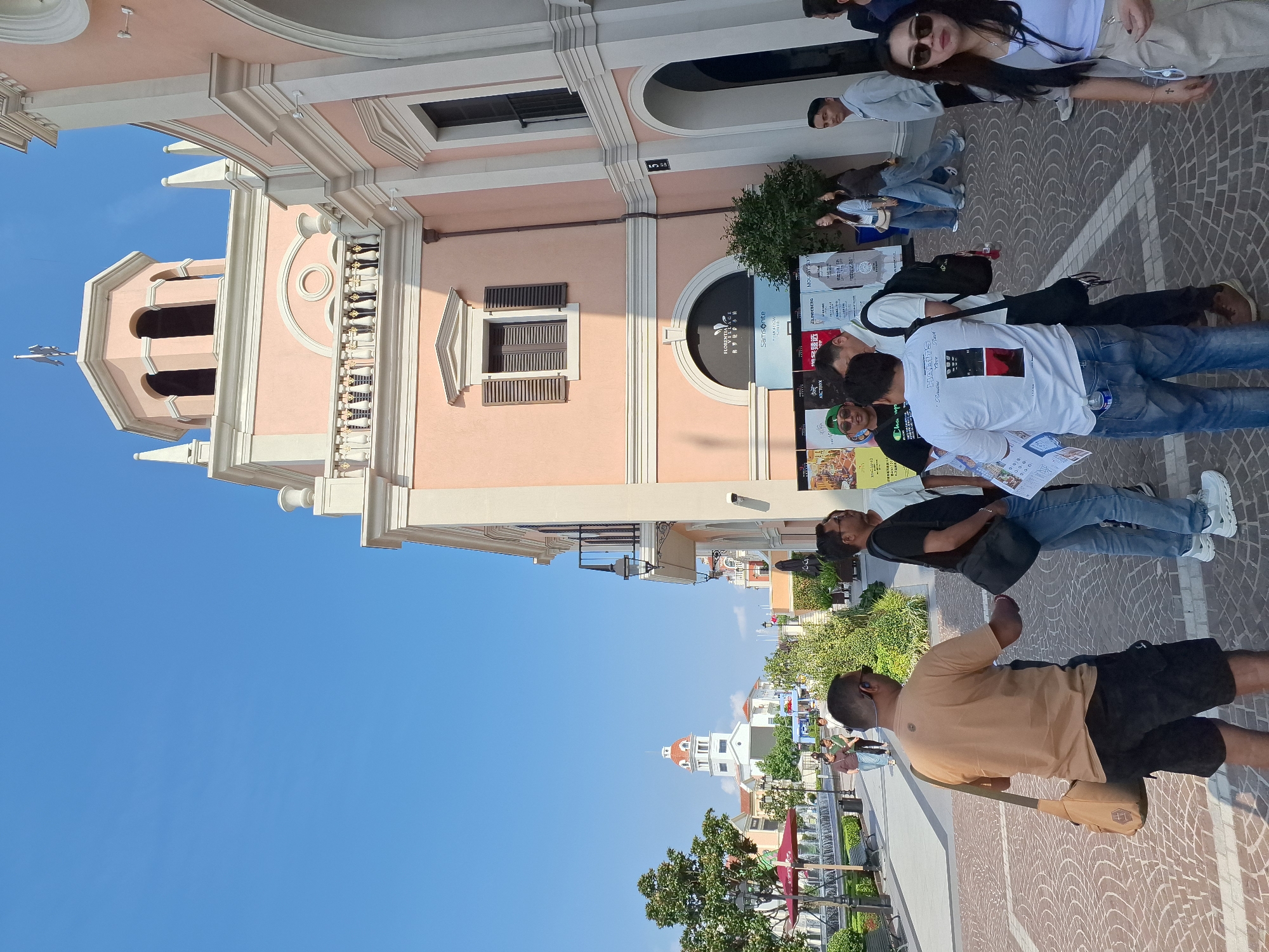The image depicts a sunny outdoor scene in the city of Pudong, China, likely at an outdoor shopping village. In the foreground, several individuals are gathered, some appearing to be tourists. A man in a tan t-shirt and black shorts is looking to his right, wearing a satchel across his body. To his left, a man in a black t-shirt and jeans is looking forward. Another man in a white t-shirt with a graphic print is holding a folded brochure.
The background features light pink buildings with architectural elements reminiscent of European styles, including arched windows, balconies, and decorative trim. A prominent clock tower with a red-domed roof is visible in the distance. The ground is paved with patterned cobblestones. Lush green trees and shrubbery are scattered throughout the area, adding to the pleasant ambiance. The sky is a clear, bright blue. Signs for "FLORENCE VILLAGE" and "SAMSONITE" are visible on one of the buildings, indicating a commercial area. A water feature and benches are also present, suggesting a public gathering space within the shopping village.