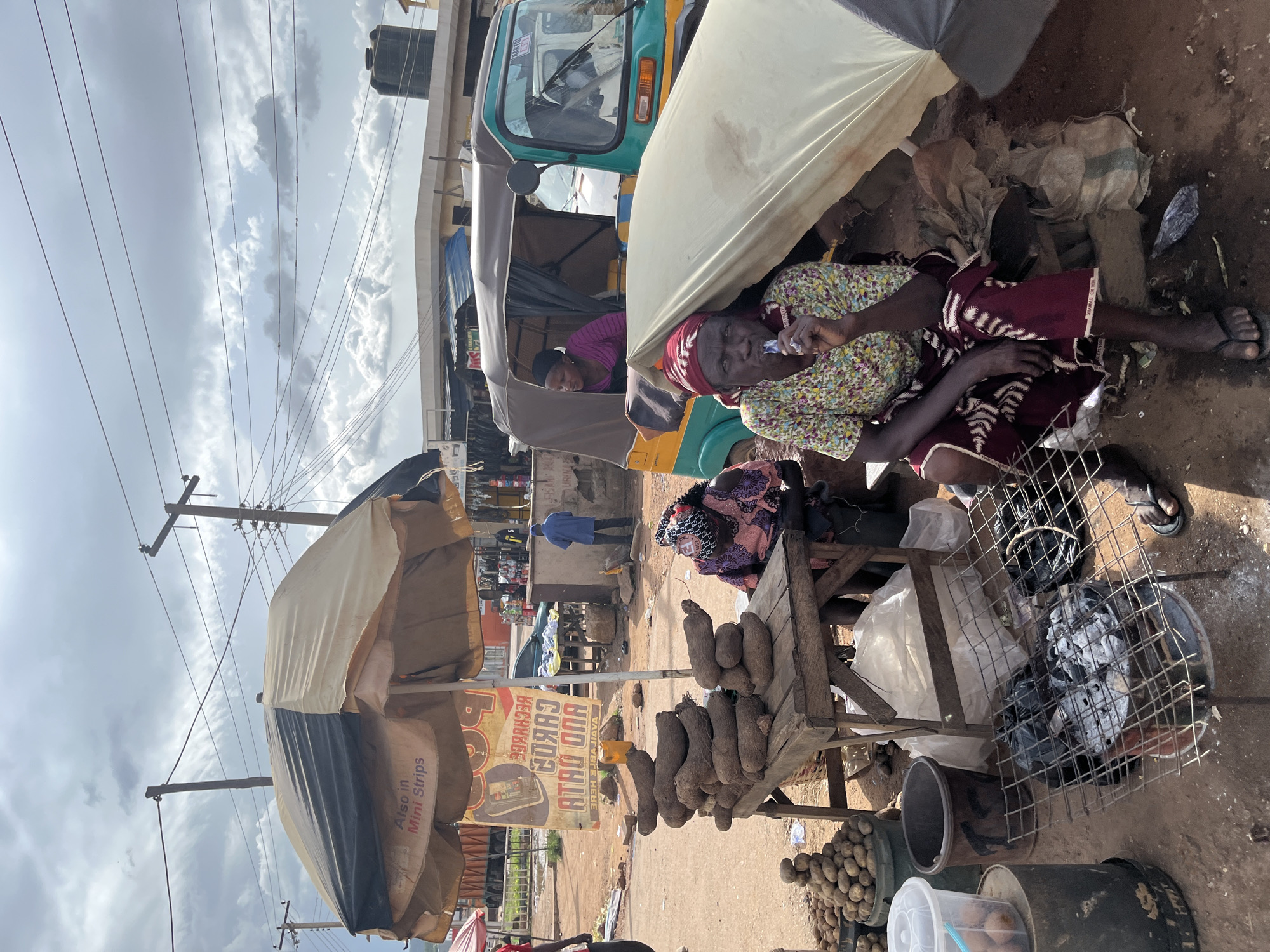 In the city of Jos, Nigeria, an outdoor market scene unfolds under a cloudy sky. A woman, wearing a headscarf and a colorful floral shirt with a patterned skirt, sits near a charcoal grill and a stack of yams. She is holding a blue object to her mouth, possibly a cigarette or a straw. Beside her, another woman in a pink shirt and headscarf is visible inside a bright yellow and green auto-rickshaw. The rickshaw is parked next to a stall covered by a large, light-colored tarp. Piles of yams are displayed on a wooden table, and a bucket of potatoes sits to the left of the frame. In the background, a large umbrella displays advertising for "Recharge Cards" and "Also in Mini Strips." Utility poles and power lines crisscross the sky, indicating an urban setting. The ground is a mixture of dirt and scattered debris. The overall impression is of a bustling market day in Jos.