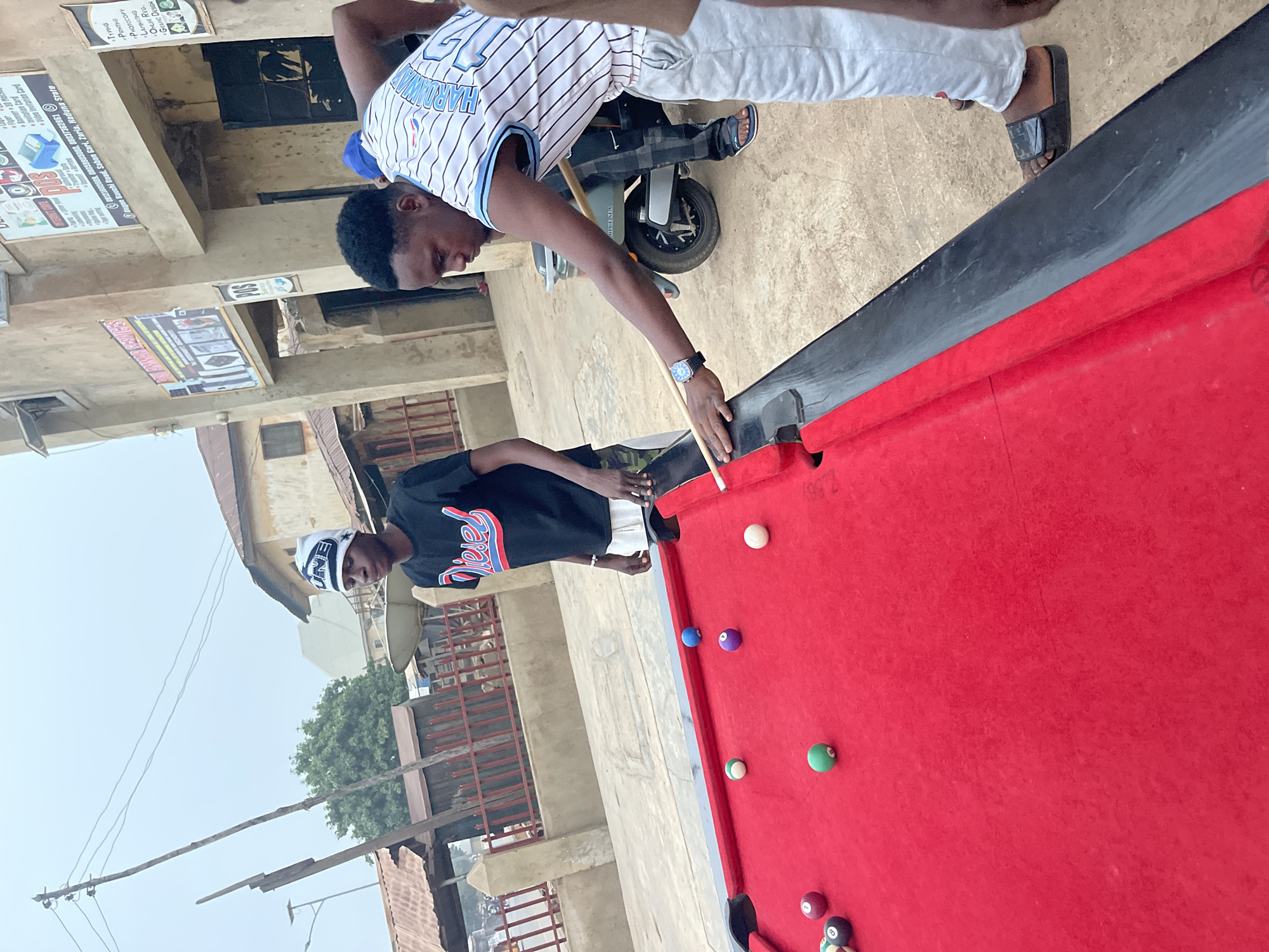 Two young men are playing pool on an outdoor table. One man is wearing a white and blue baseball jersey, white pants, and sandals. He is leaning over the table, cue in hand, taking a shot. The other man is wearing a black shirt and white beanie, standing near the table's edge. The pool table is red with various colored balls scattered across its surface. Behind the men, a scooter is parked. The scene takes place in Unguwar Gabas, Nigeria, as indicated by the signage on a nearby building. The building's signage advertises various services, including typing, printing, photocopying, laminating, ONIE registration, and graphic design. Power lines and other buildings are visible in the background. The day appears overcast.