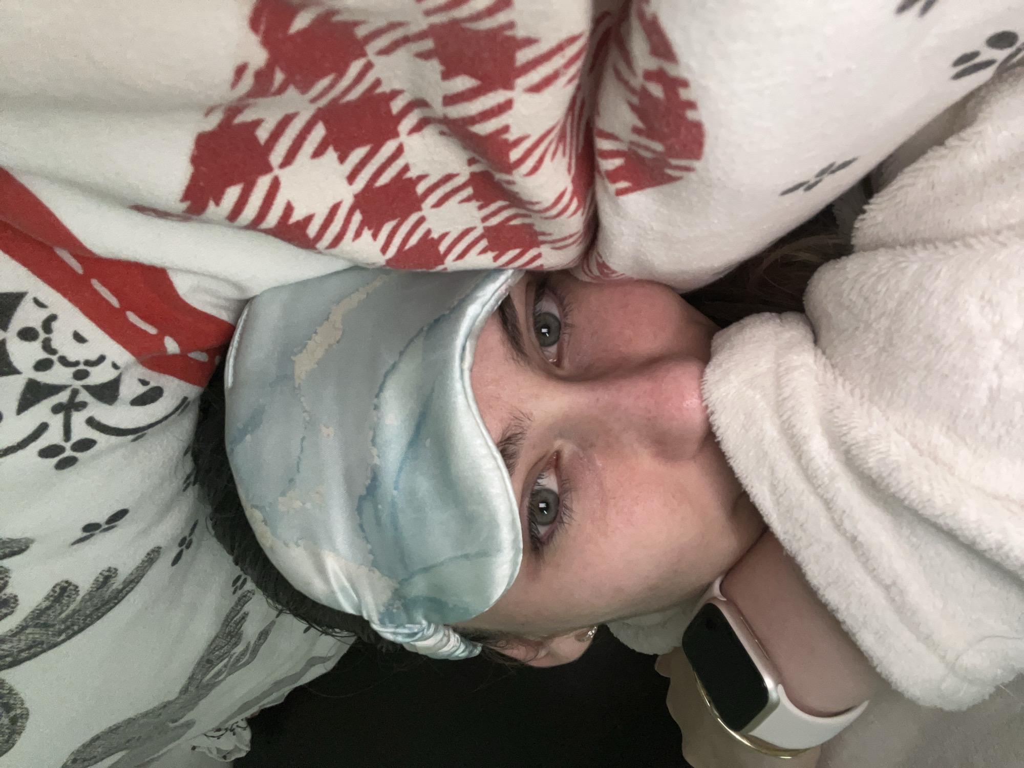A close-up, rotated portrait shot depicts a person lying in bed, covered in blankets. The person wears a light blue satin sleep mask. One blanket features a red and white houndstooth pattern, while another is white with gray cactus designs. A fluffy white blanket or robe also appears. The person's face is visible, showing gray eyes and fair skin. A white smartwatch with a gold band is on the person's wrist. The location is Unknown, Unknown.