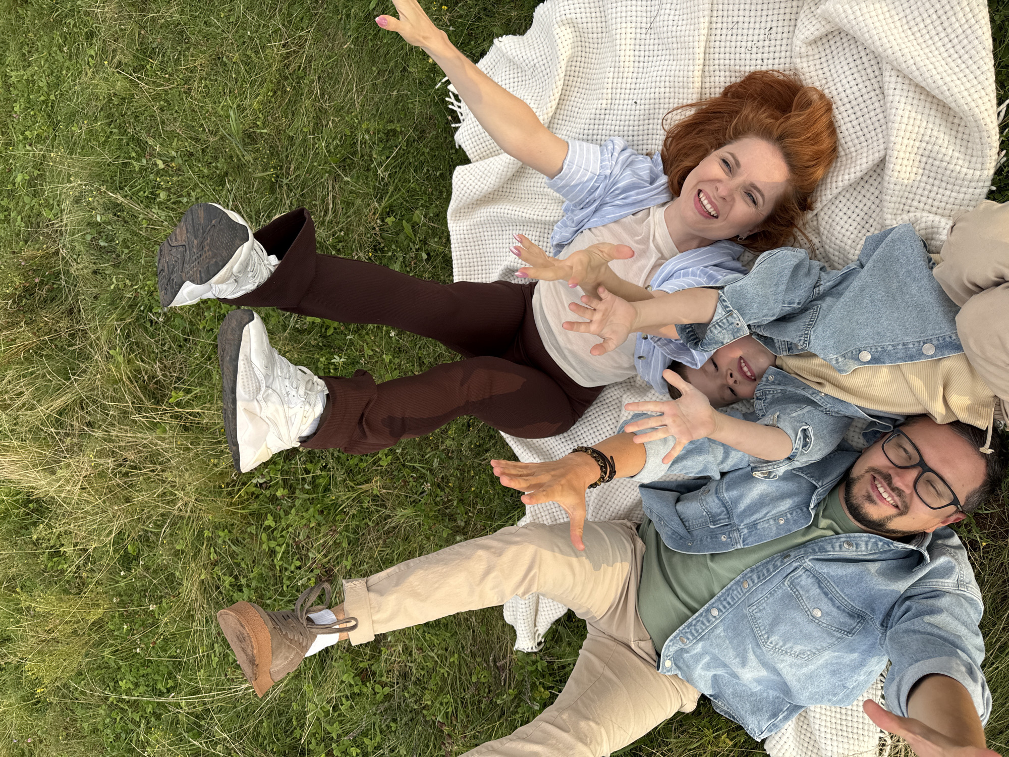 A family of four is lying on a picnic blanket in a grassy field. The parents are lying on their backs with their legs raised in the air, while their two young children are lying on top of them. The family is smiling and laughing, and they appear to be enjoying their time together. The location is a grassy field outside of Olkhovka, Russia. The weather appears to be warm and sunny.